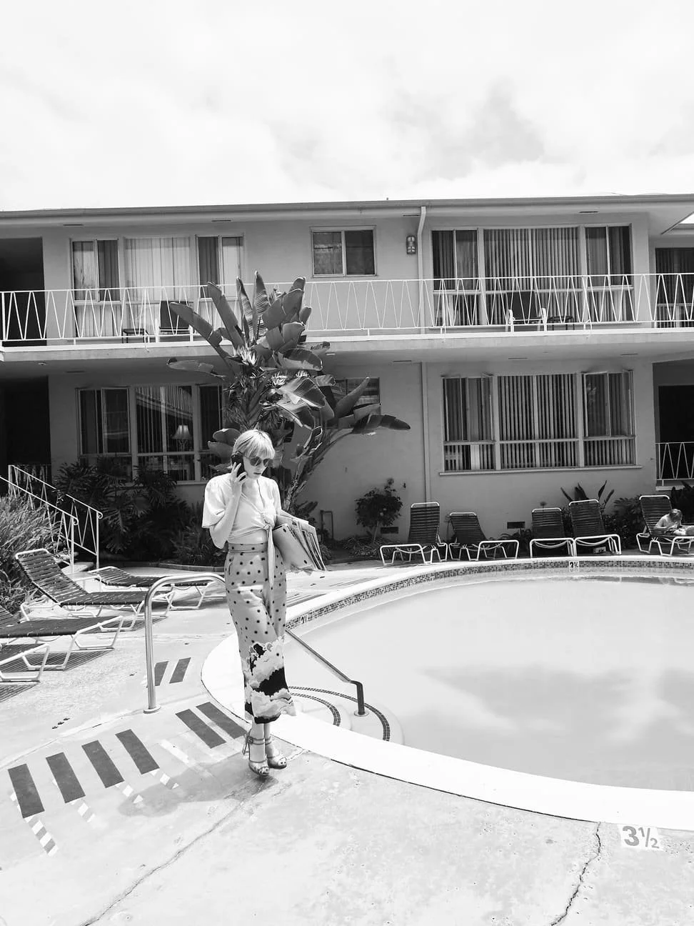 Een vrouw met een koptelefoon, zonnebril en een lange, polkadot jurk staat naast een zwembad en leest een tijdschrift. Op de achtergrond zijn ligstoelen, planten en een appartementencomplex te zien gefotografeerd door Eric van den Elsen.