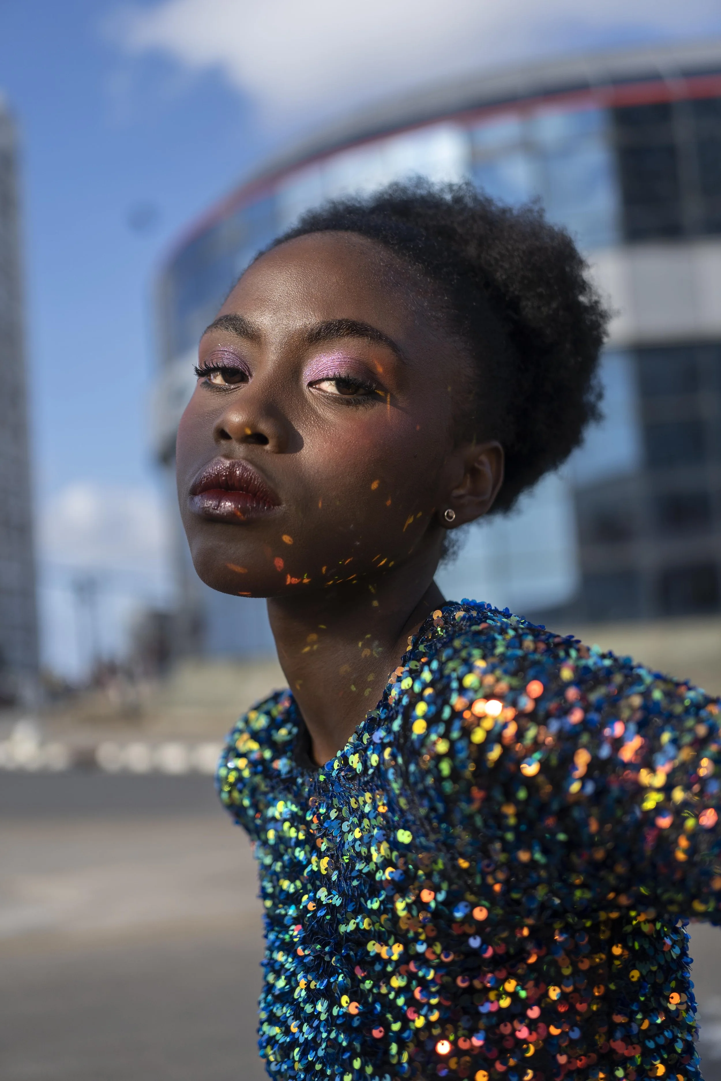 Portret van een vrouw met donkere huid, kleurrijke oogmake-up, en een glanzende, gekleurde sequin jurk, op een stedelijke achtergrond onder een blauwe lucht gefotografeerd door Eric van den Elsen.