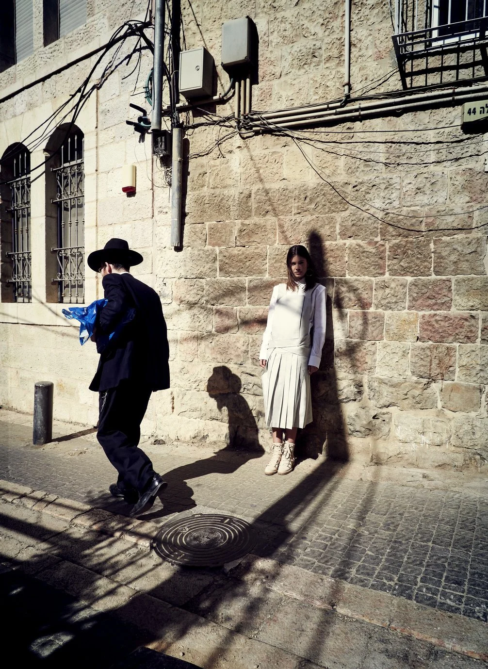 Een vrouw leunt tegen een stenen muur, terwijl een man in een hoed verderop loopt op de stoep. Het is zonnig, en er zijn kabels en elektriciteitskasten op de muur naast hen gefotografeerd door Eric van den Elsen.