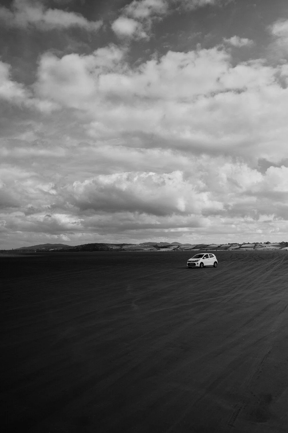 Een witte auto rijdt op een uitgestrekt, vlak landschap met een bewolkte hemel in het zwart-wit.