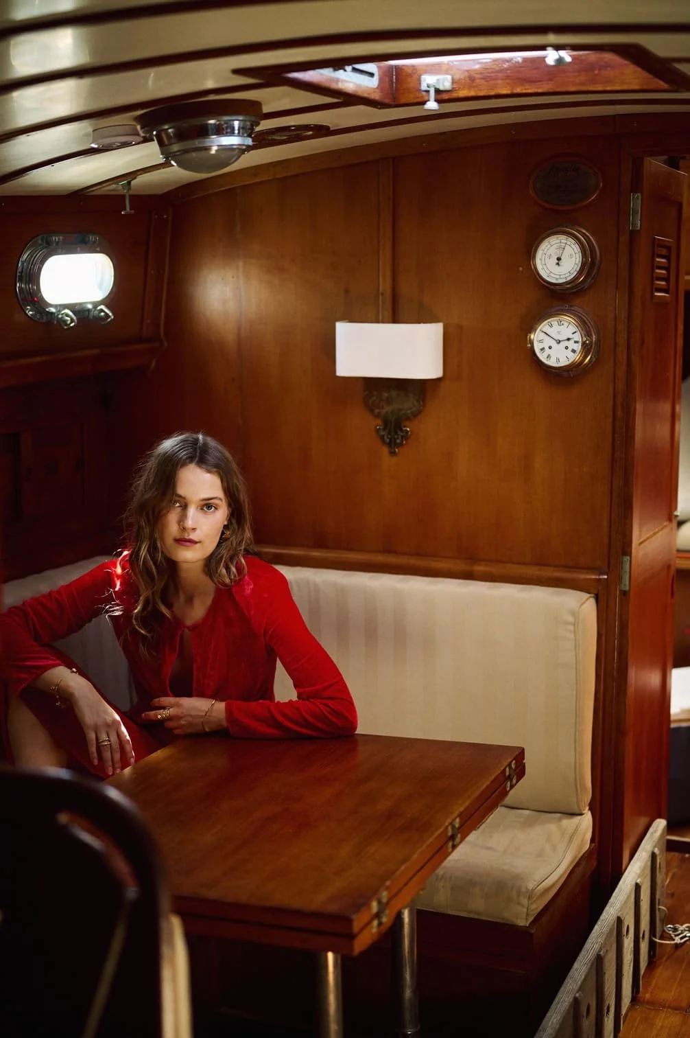 Een vrouw in een rood jasje zit aan een houten tafel in een houten interieur van een schip, met vintage klokken en een lamp aan de muur.