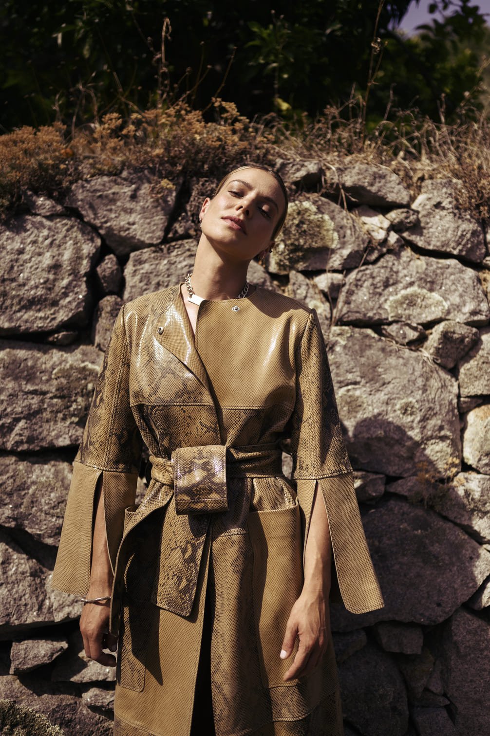 Sallie Harmsen staat voor een stenen muur, draagt een glanzende, beige, snake-achtig patroon jas met ceintuur, haar hoofd licht omhoog en de zon schijnt op haar gezicht gefotografeerd door Eric van den Elsen.