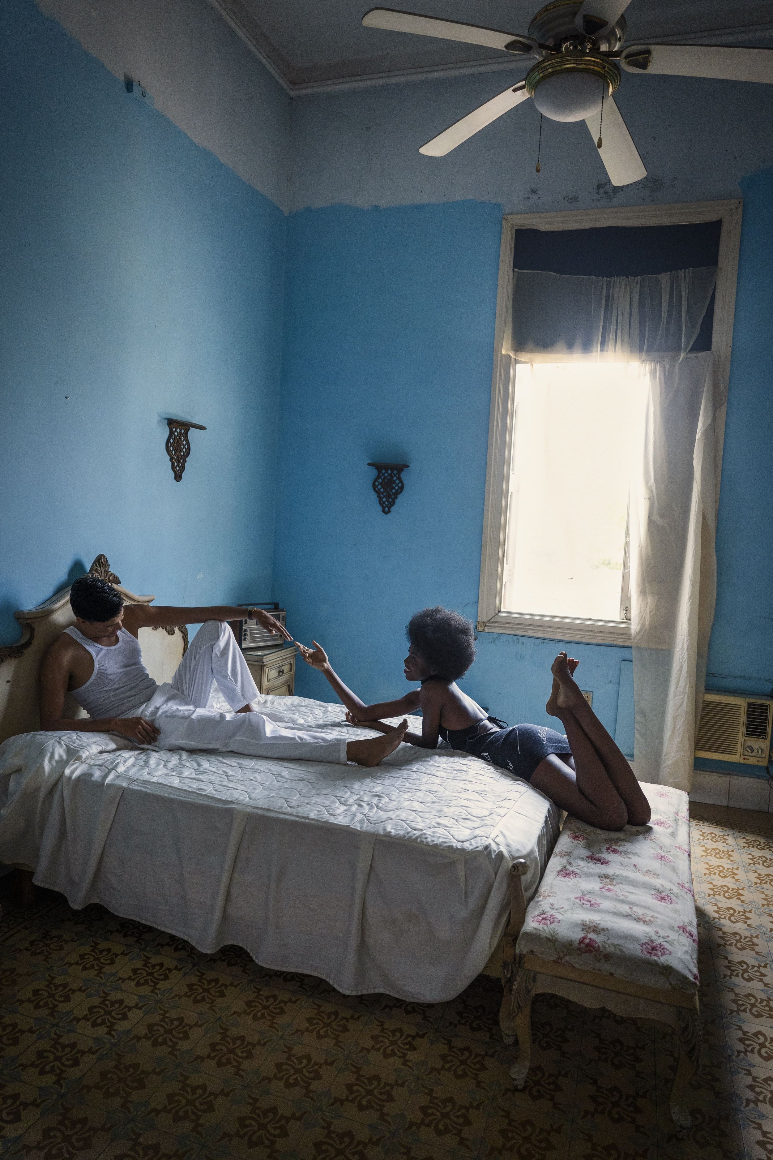 Een slaapkamer met blauwe muren, een bed met een man en een vrouw. De man ligt op het bed en de vrouw knielt naast hem, beiden lijken te praten gefotografeerd door Eric van den Elsen.