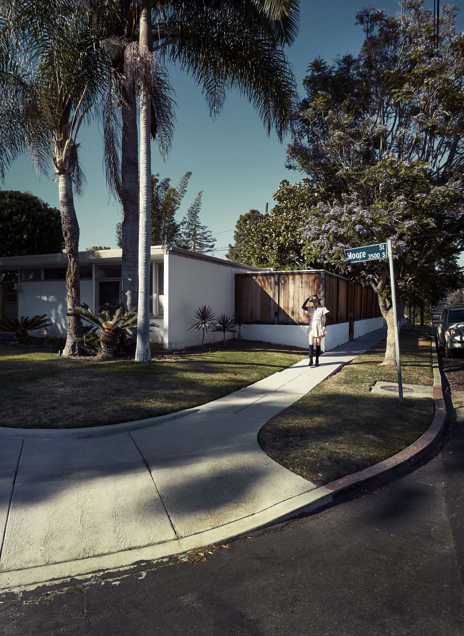 Een straataanzicht met een huis, palm- en andere bomen, een voetpad, een vrouw met hoed en laarzen die door de straat loopt gefotografeerd door Eric van den Elsen.