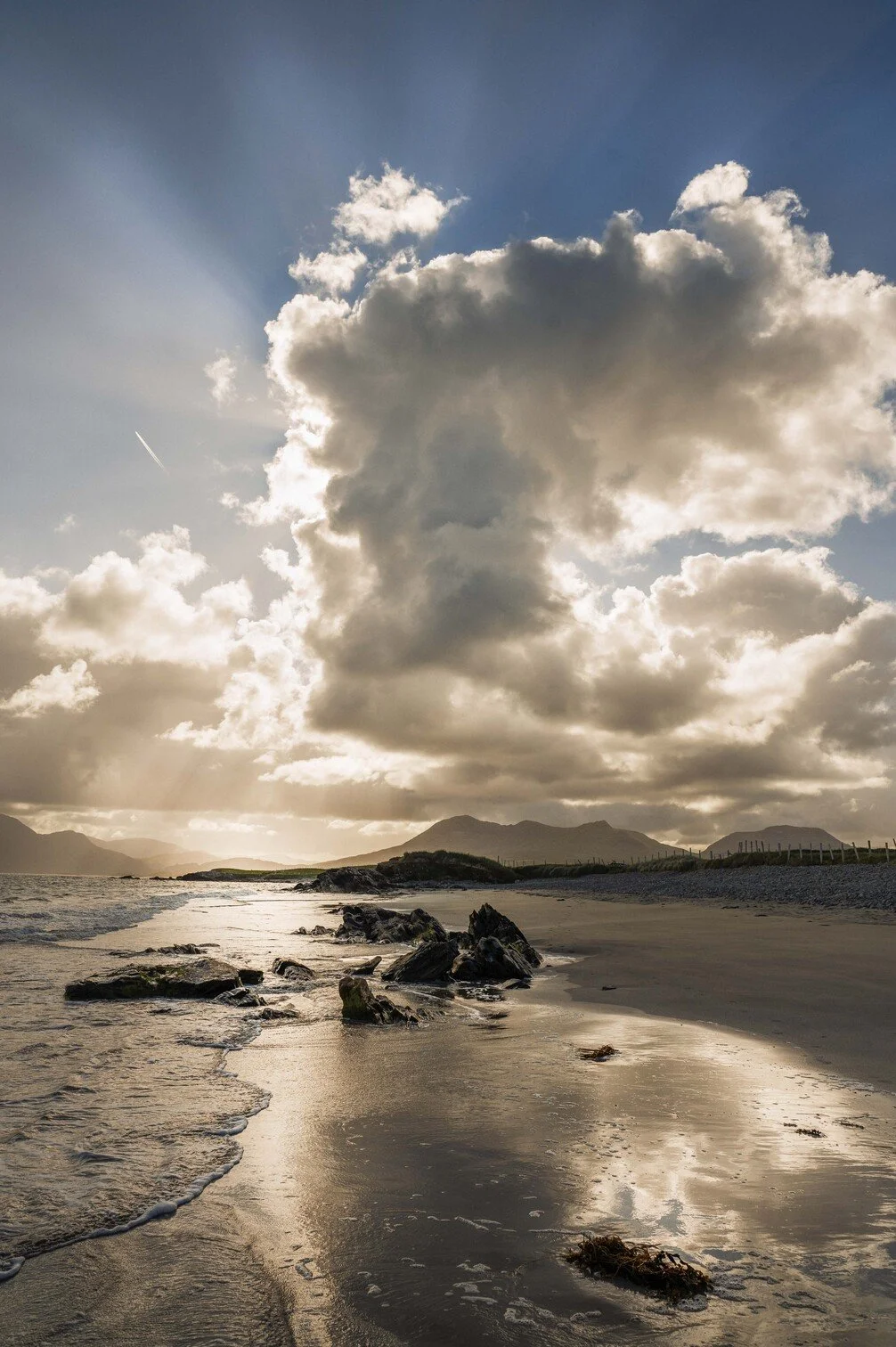 Een rustige kustlijn met rotsen, zandstrand en een bewolkte hemel met zonnestralen die door de wolken breken, met bergen op de achtergrond gefotografeerd door Eric van den Elsen.