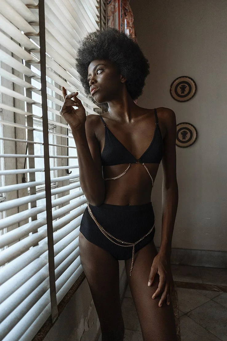 Een vrouw met een grote, natuurlijke afro-haar staat naast een raam met luiken en kijkt buiten gefotografeerd door Eric van den Elsen.