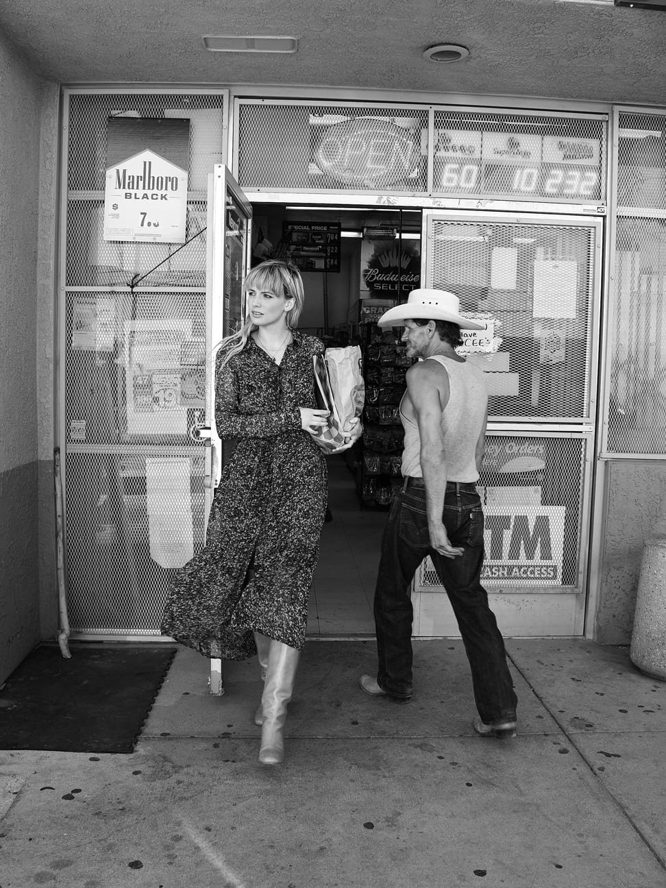 Een vrouw en een man staan bij de ingang van een convenience winkel. De vrouw houdt een zak en een boek of tijdschrift vast en kijkt naar links. De man draagt een hoed en een zonder mouwen shirt, en staat met zijn hand in zijn broekzak gefotografeerd