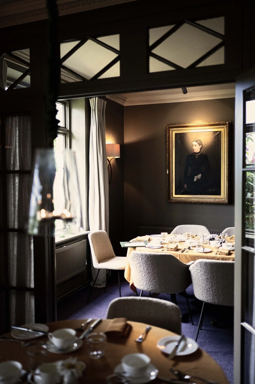 Een elegant, gedekte eethoek met een schilderij aan de muur, grote ramen met witte gordijnen, en een ronde tafel met wit servies en bestek, in een sfeervolle kamer gefotografeerd door Eric van den Elsen.