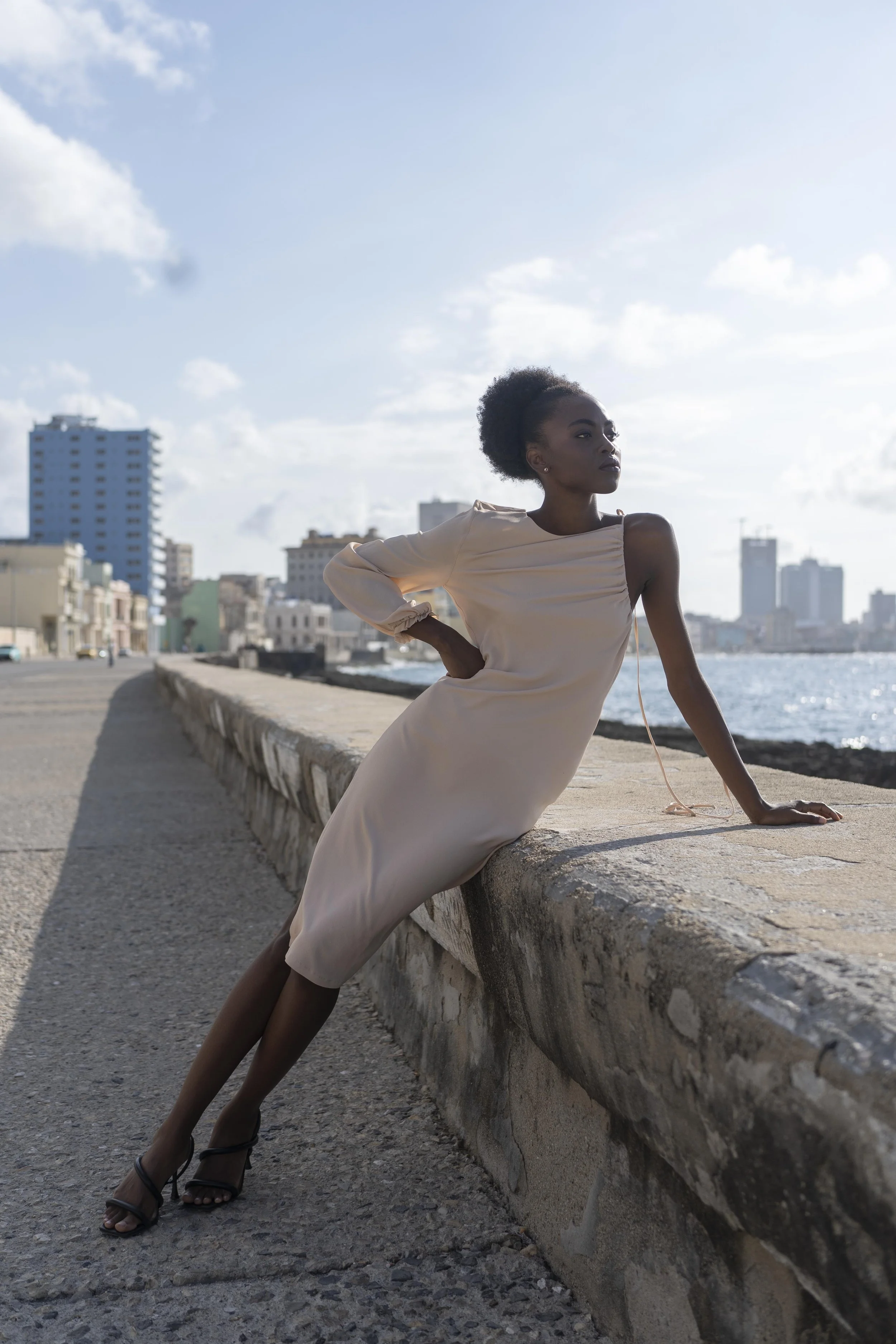 Een vrouw in een beige jurk poseert langs de boulevard met de stad en de oceaan op de achtergrond gefotografeerd door Eric van den Elsen.
