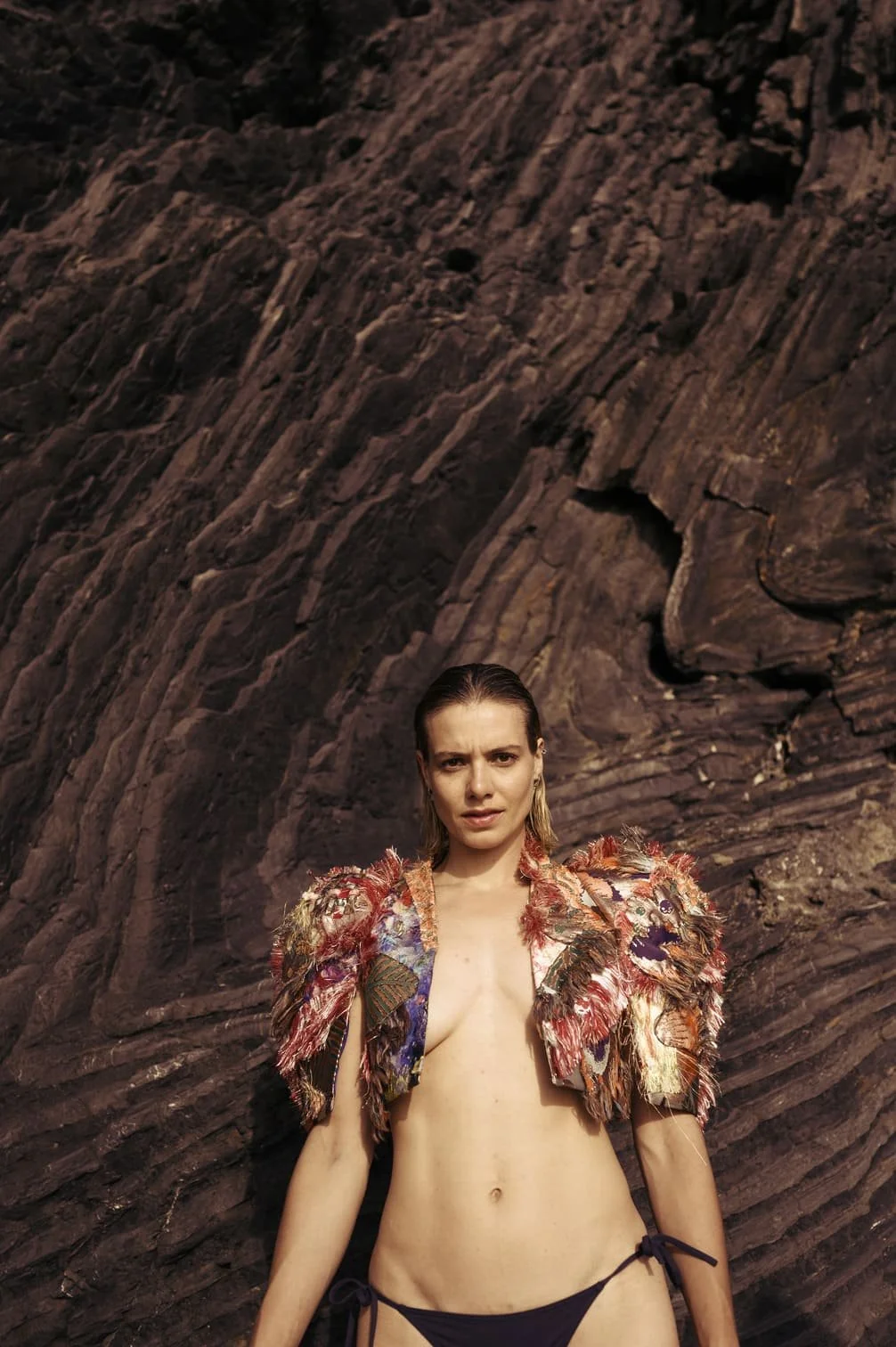 Sallie Harmsen met nat haar, gekleed in een kleurrijk, bontachtige jas die open is en geen top draagt, staat voor een rotsachtige achtergrond gefotografeerd door Eric van den Elsen.