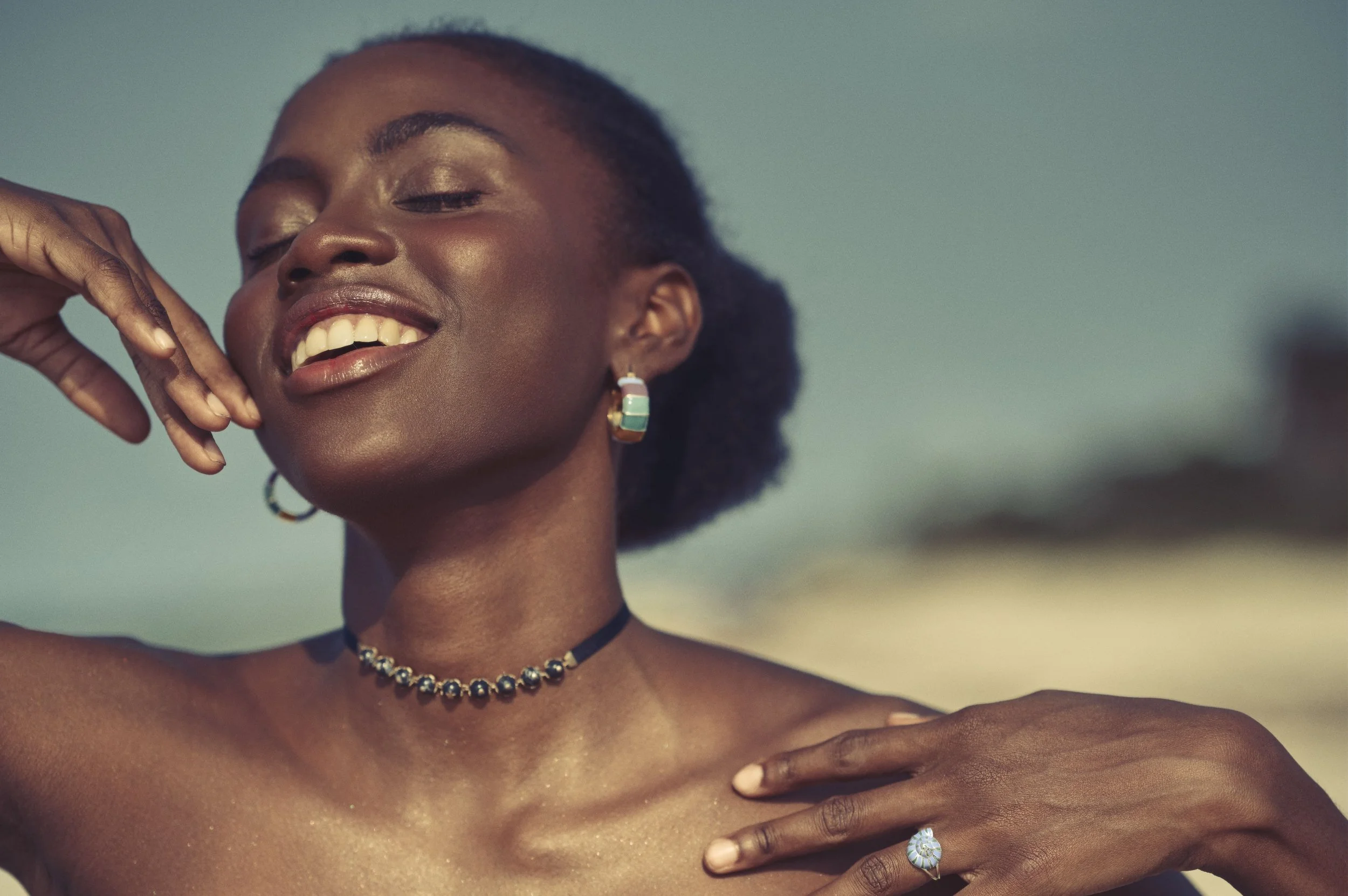 Portret van een lachende Cubaanse vrouw met donkere huid, sieraden en glanzende huid, met een blauwe lucht op de achtergrond gefotografeerd door Eric van den Elsen.