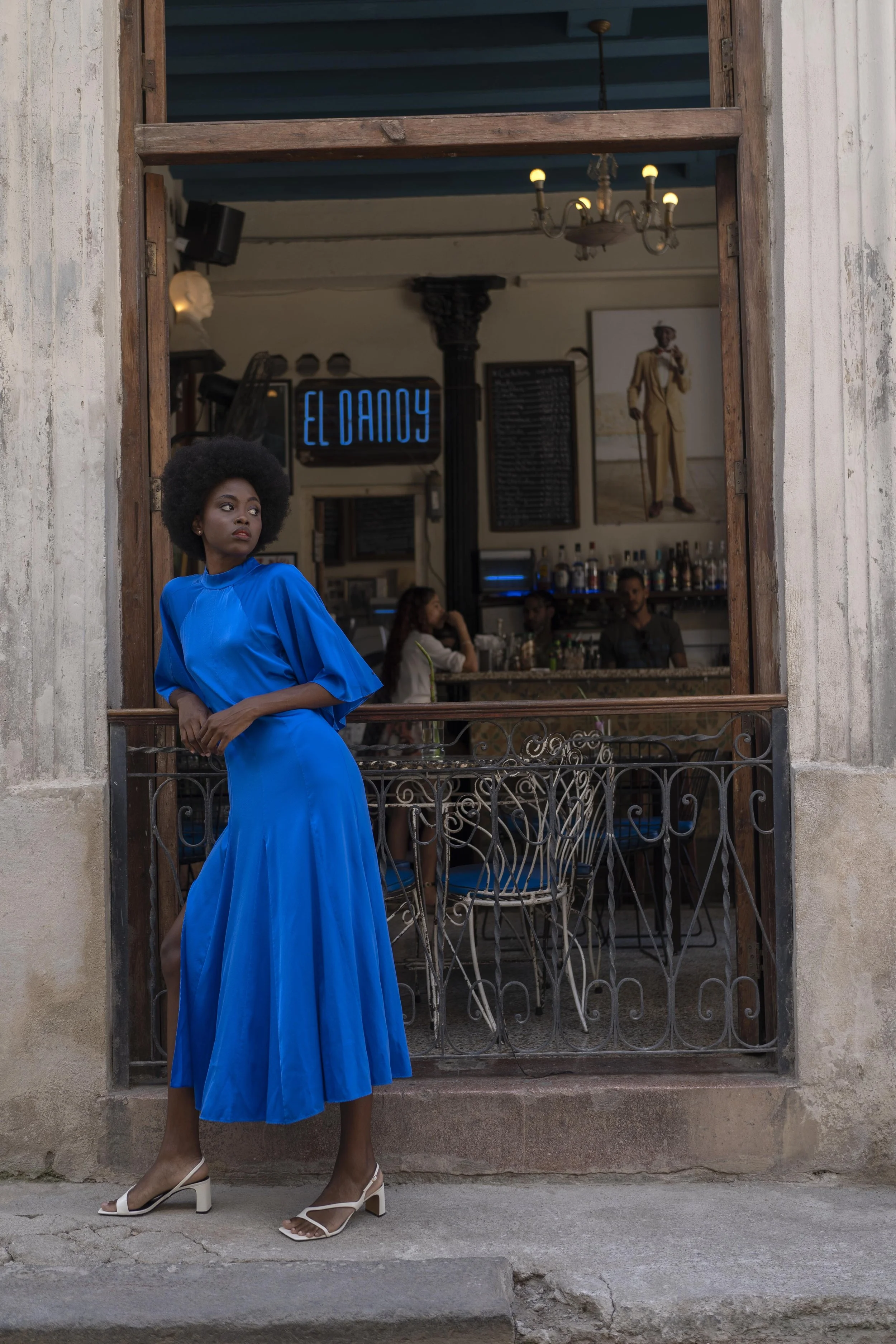 Een vrouw in een felblauwe jurk staat op een stoep voor een café met een open raam. Achter het raam is een bar te zien met klanten, en een neonbord dat 'El Dancy' leest  gefotografeerd door Eric van den Elsen.