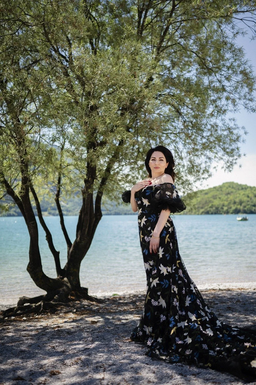 Nazmiye Oral in een lange, zwarte jurk met bloemenpatroon staat op een strand bij een water, onder een boom, met een platteland op de achtergrond gefotografeerd door Eric van den Elsen.