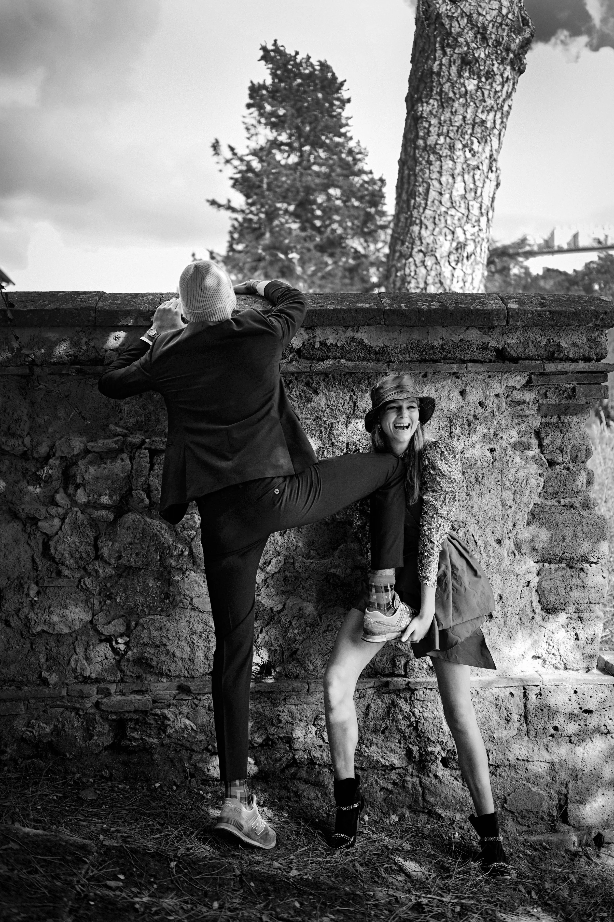 Twee mensen die humoristisch in een steegje poseren: een man die probeert te 'trappen' naar een lachende vrouw, terwijl ze zich onder een stenen muur houdt. Het is een zwart-wit foto gefotografeerd door Eric van den Elsen.