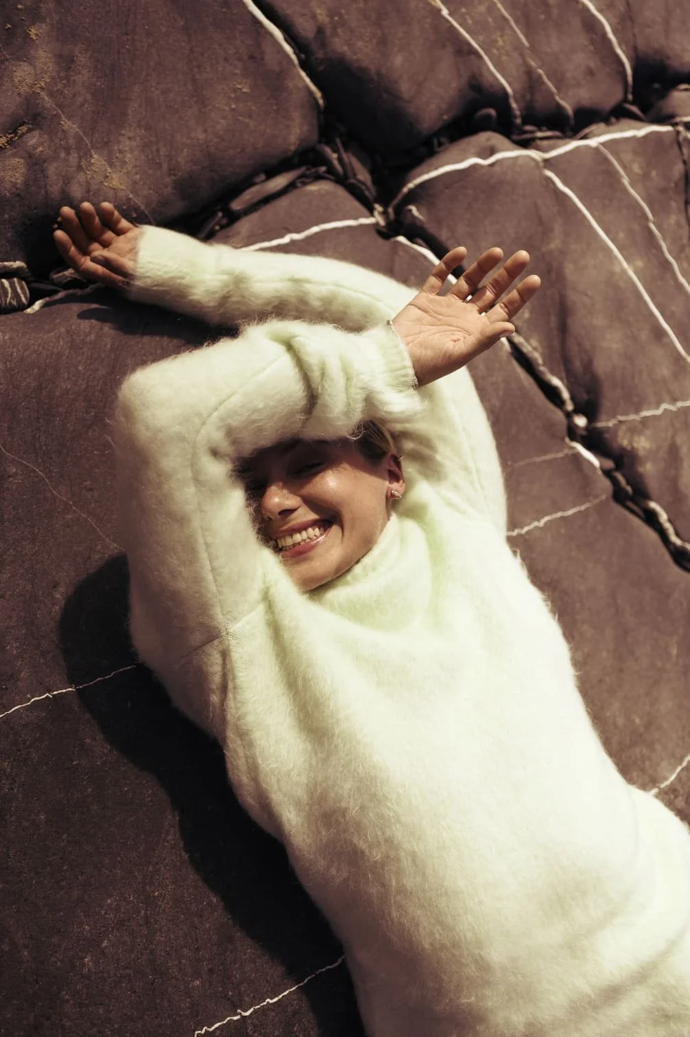 Een lachende Sallie Harmsen ligt op een bank, gekleed in een witte trui, met haar armen over haar hoofd en haar ogen dicht gefotografeerd door Eric van den Elsen.