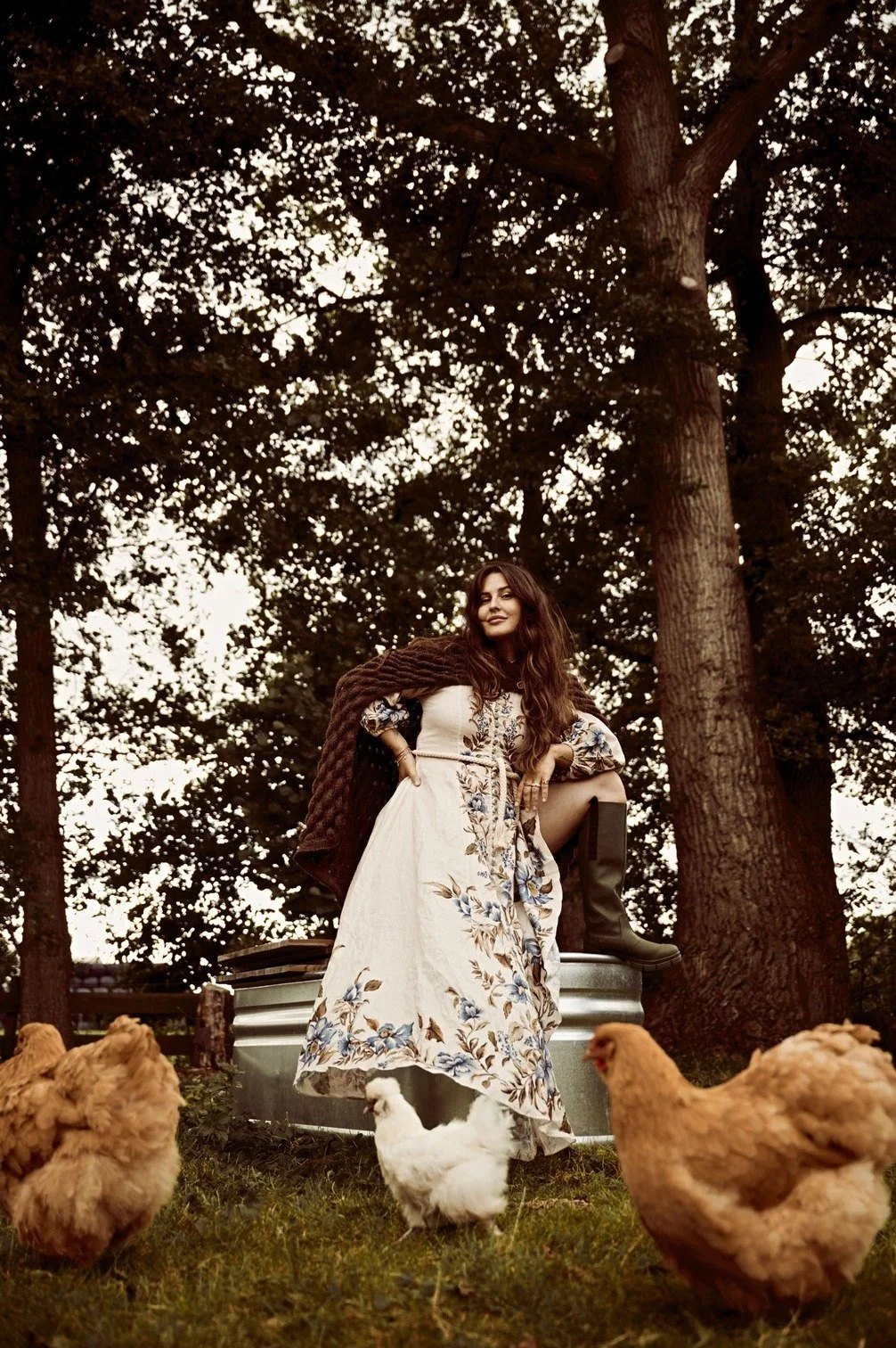 Romy Boomsma poseert in een bos met drie kippen op de voorgrond, draagt een lange, bloemenprint jurk en laarzen, met een warme deken over haar schouders gefotografeerd door Eric van den Elsen.