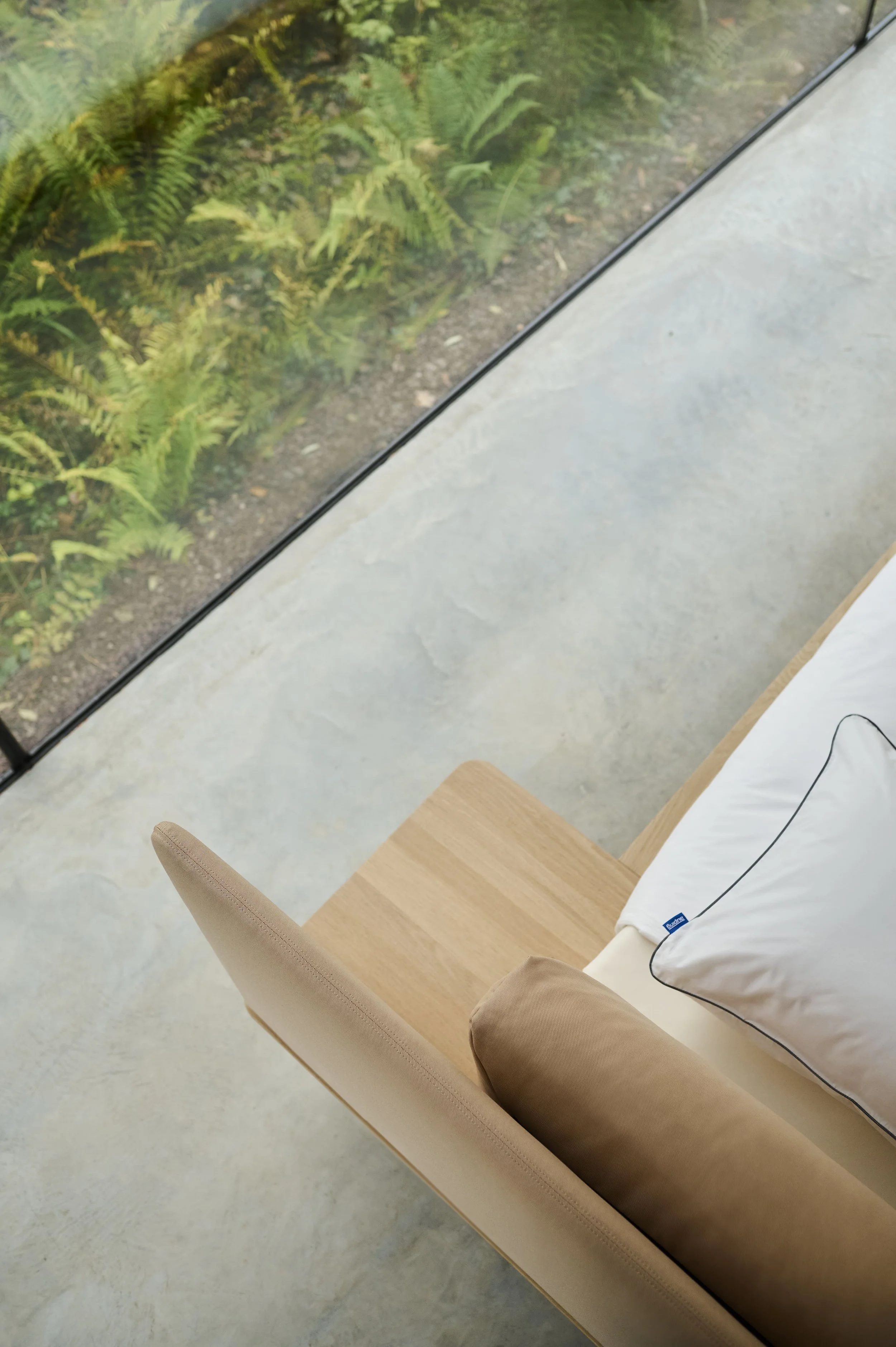 Uitzicht vanuit een kamer met groot raam op een groene tuin met farn en planten, naast een Auping bed met witte beddengoed en een beige kussen gefotografeerd door Eric van den Elsen voor Auping.