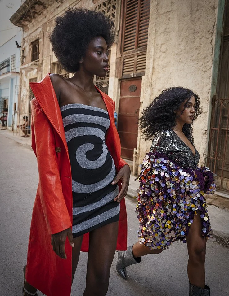 Twee vrouwen wandelen langs een oude stenen muur in een stad, een in een zwart-grijs gestreept jurk en een in een kleurrijke sequin jas gefotografeerd door Eric van den Elsen.