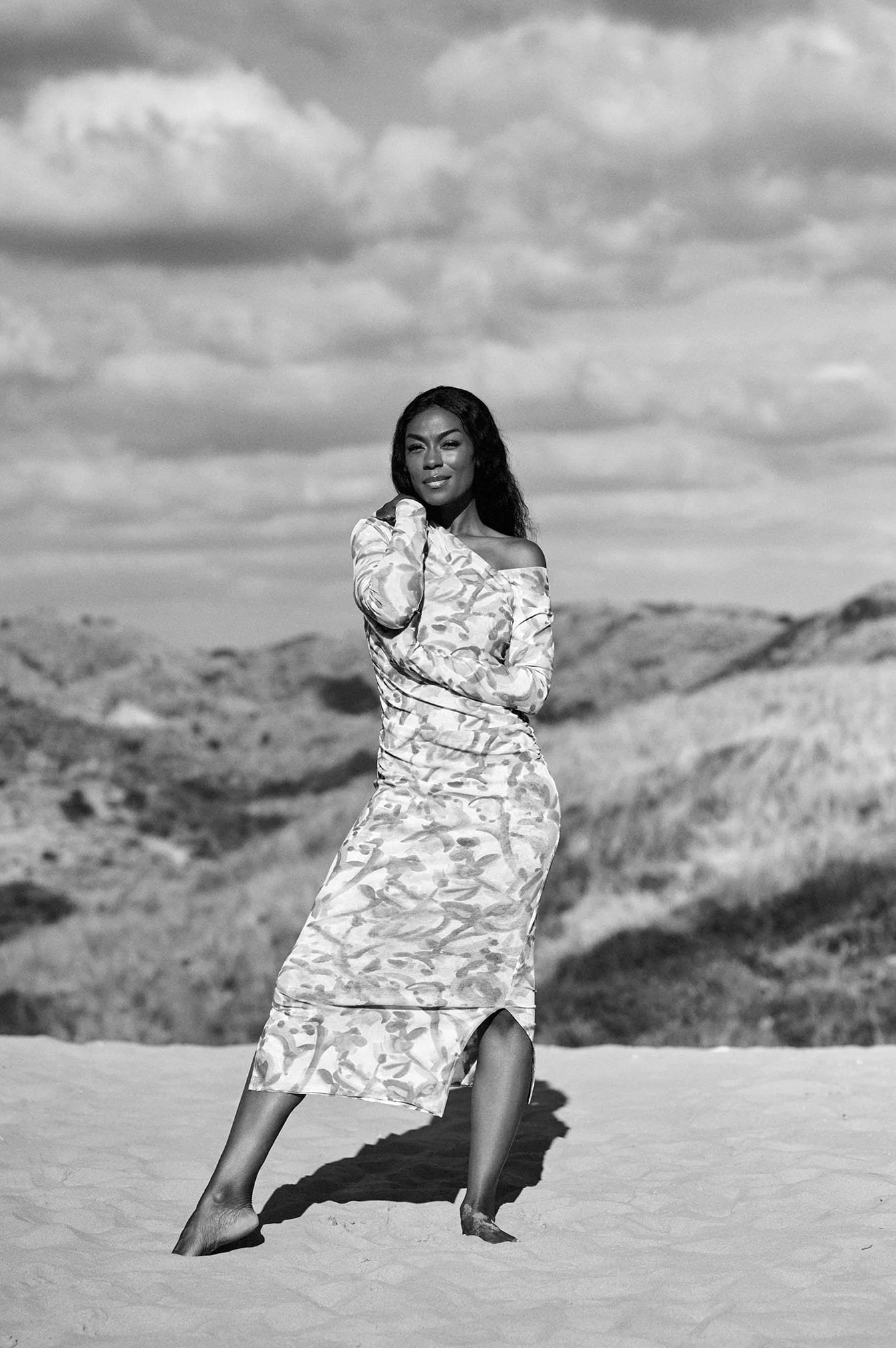 Giovanca Ostiana poseert op zand, met een landschap en wolken op de achtergrond, in zwart-wit gefotografeerd door Eric van den Elsen.