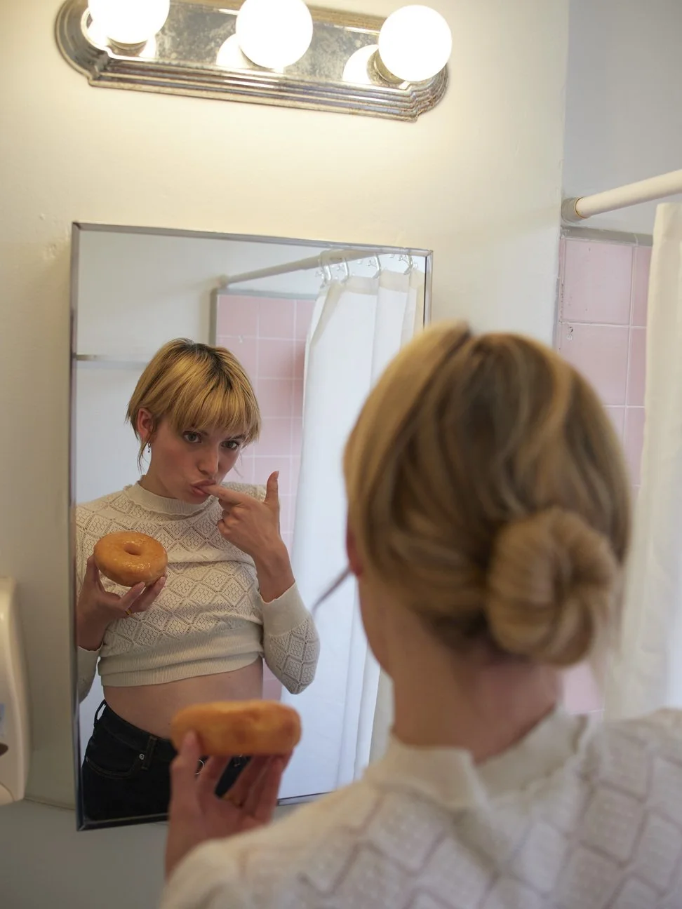 Een jonge vrouw kijkt in de spiegel terwijl ze twee donuts vasthoudt, één in elke hand. Ze raakt haar lip aan met haar vinger en poseert speels gefotografeerd door Eric van den Elsen.