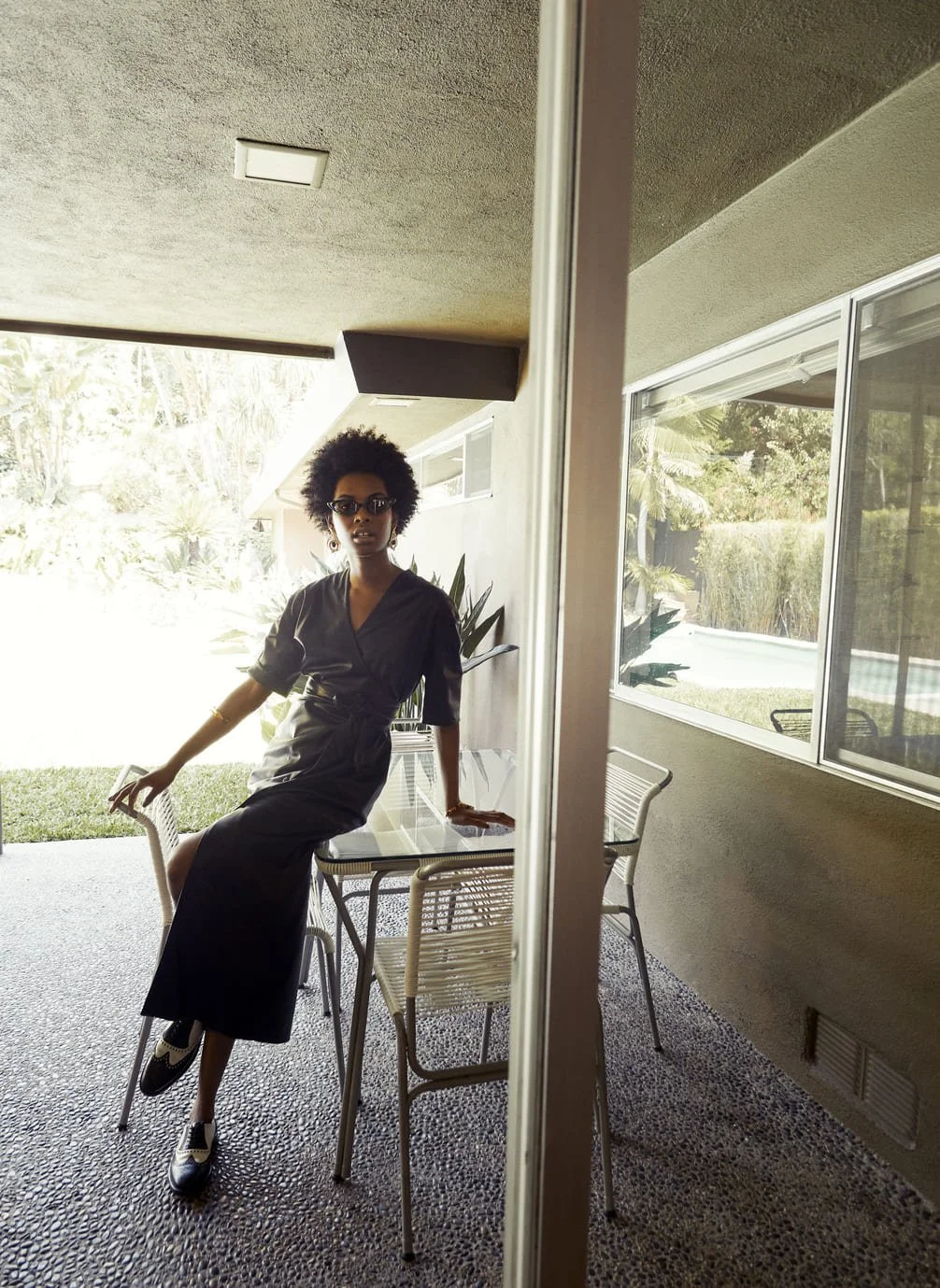 Een vrouw met kort krullend haar zit buiten op een glaskeramisch tafel met vier stoelen. Ze draagt een lange zwarte jurk, sunglasses en zwart-witte schoenen. Er is een tuin met groen en planten op de achtergrond gefotografeerd door Eric van den Elsen