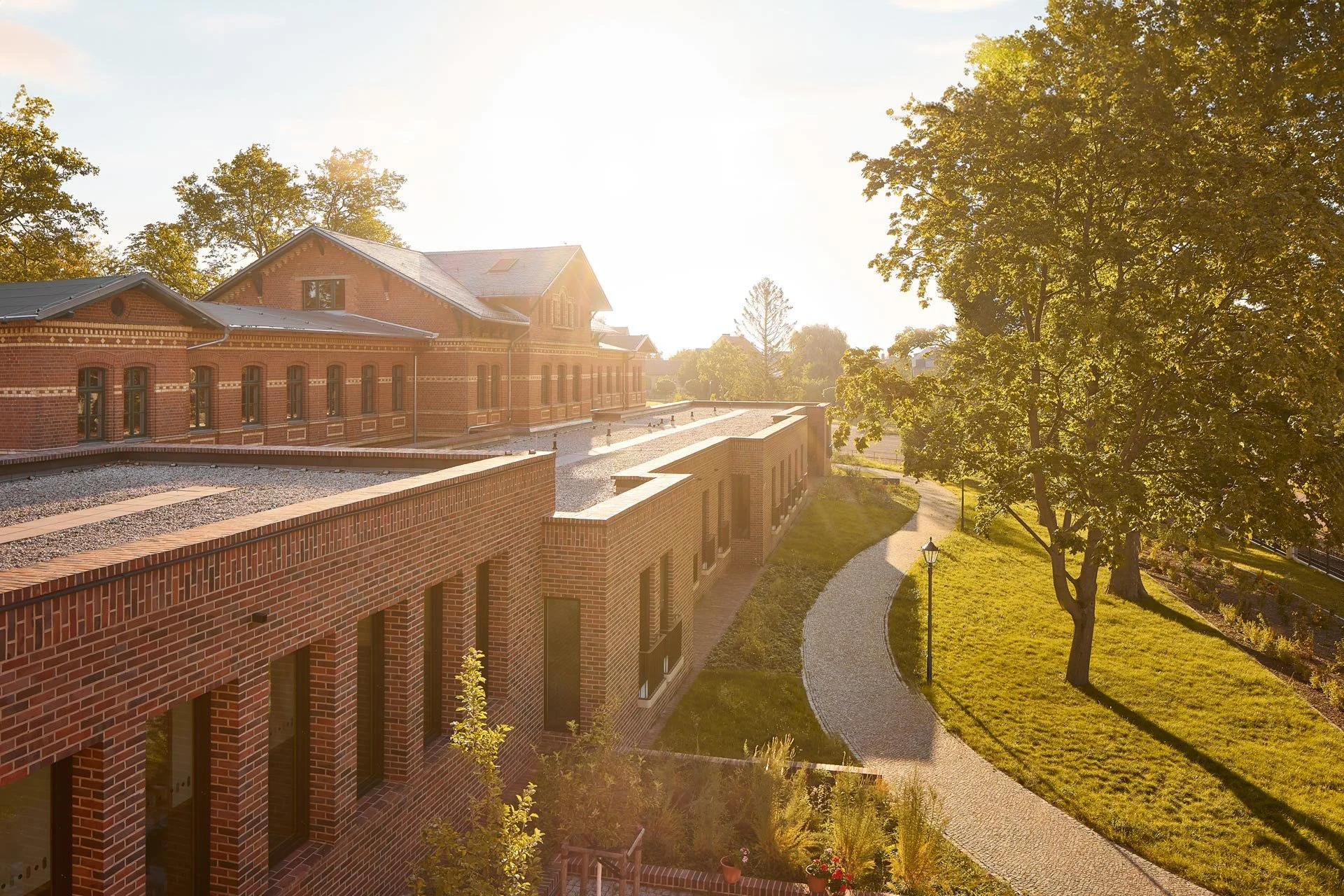 Backsteinbau mit Fenstern in einem parkähnlichen Umfeld, Bäume und Rasenfläche bei Sonnenuntergang.