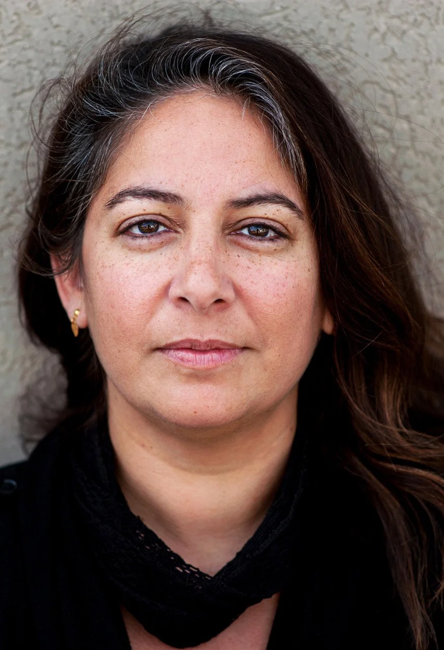 Close-up of a woman with brown hair, light skin, and brown eyes, wearing a black top and gold earring, posing against a beige textured background.