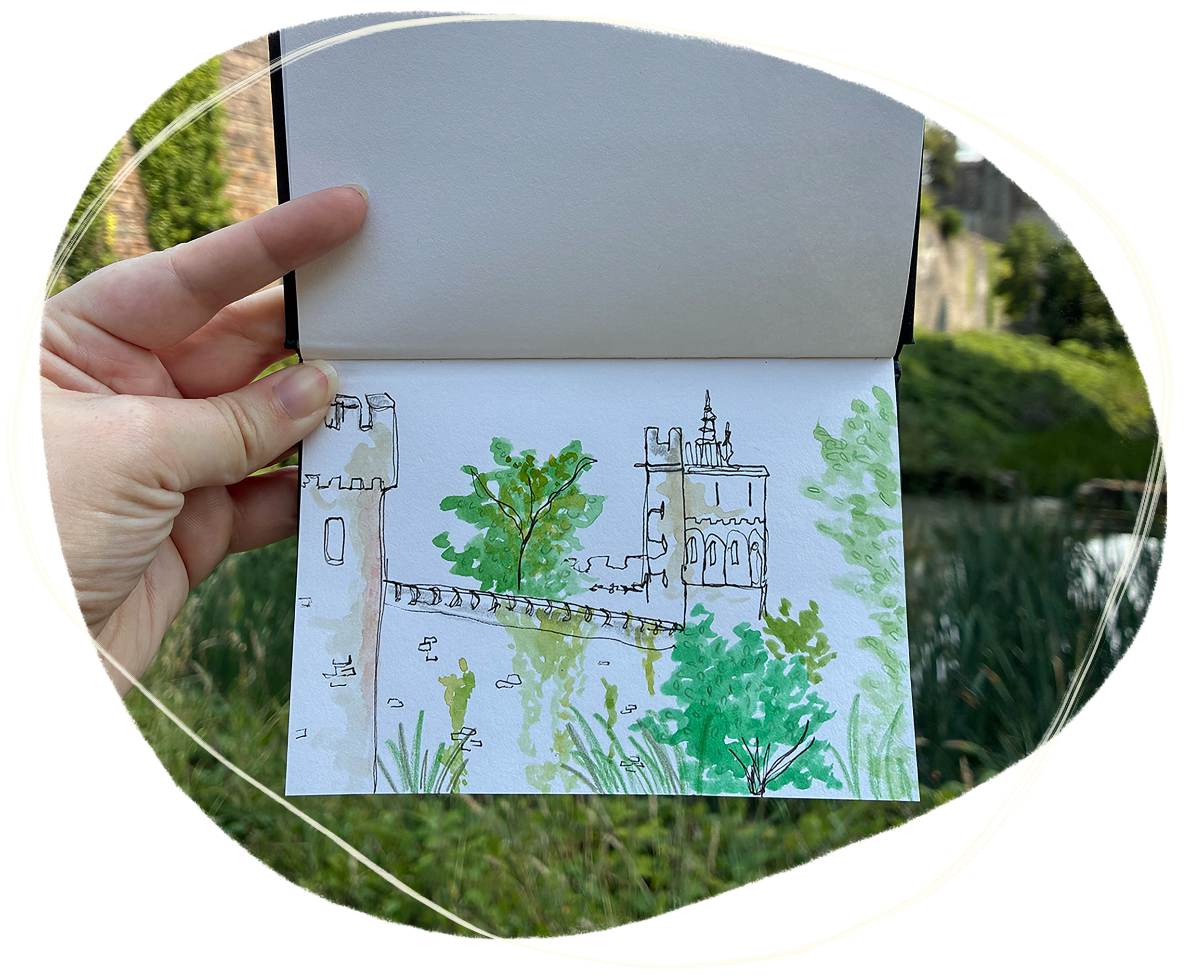 Hand holding a sketchbook of a watercolour sketch of Cardiff castle, with Cardiff castle seen in the background of the image