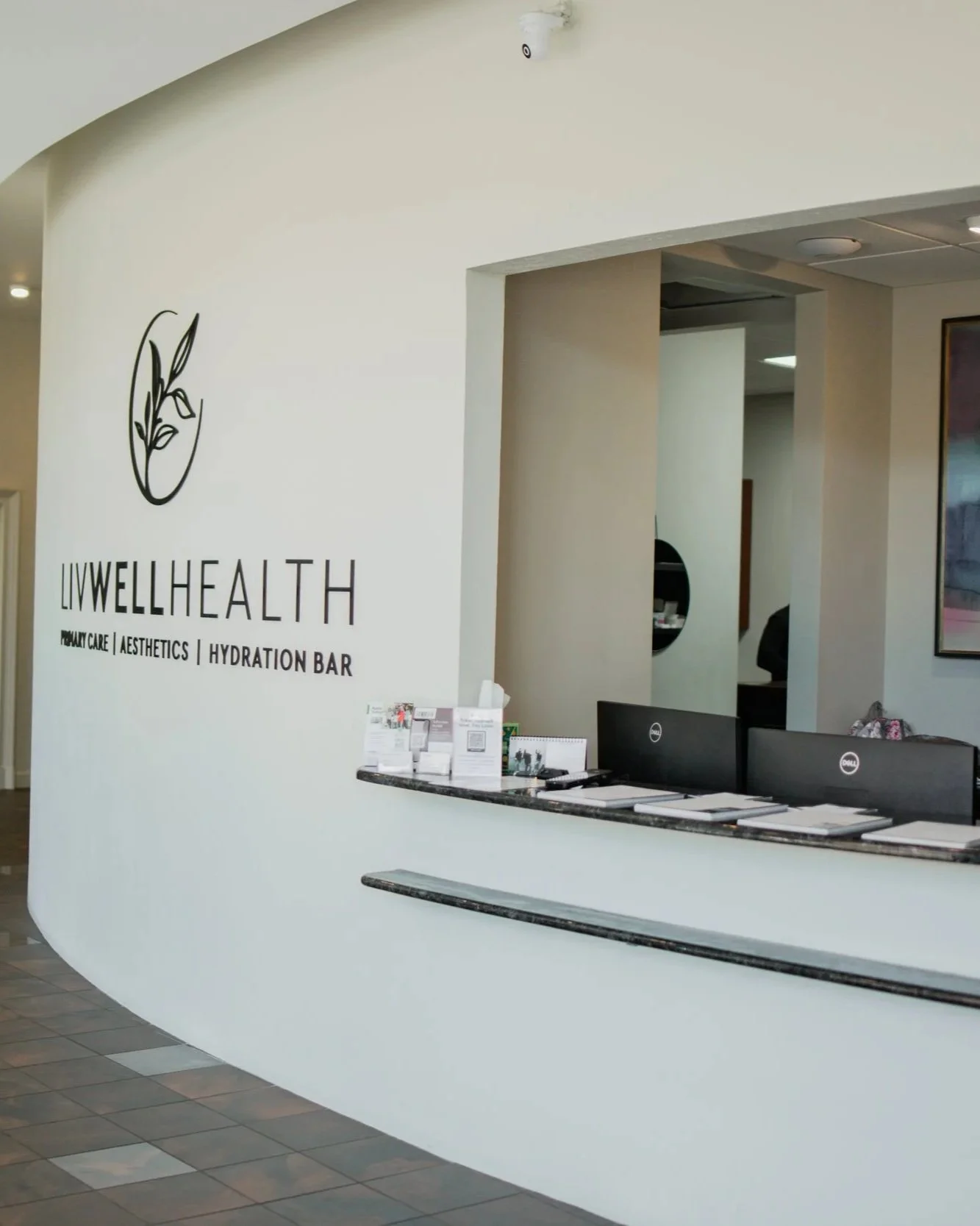 Reception area of a health clinic with a white curved wall displaying a logo and text that reads 'LiveWell Health' with services listed below. A black marble counter with brochures and two computer monitors is in front of the wall. The background includes an opening leading to another room with a mirror on the wall.