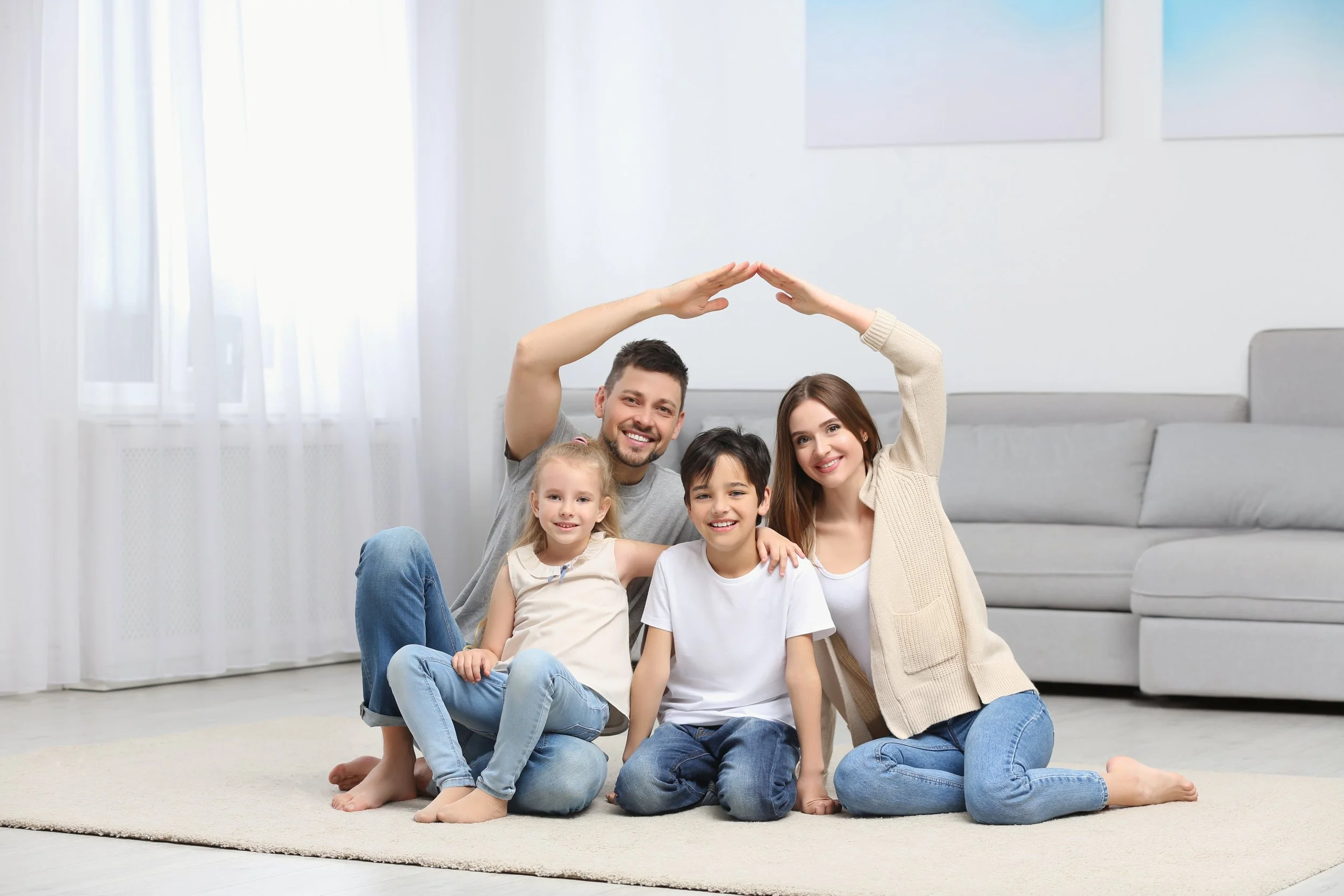 Happy family posing on carpet in living room, parents forming roof shape with arms over children.