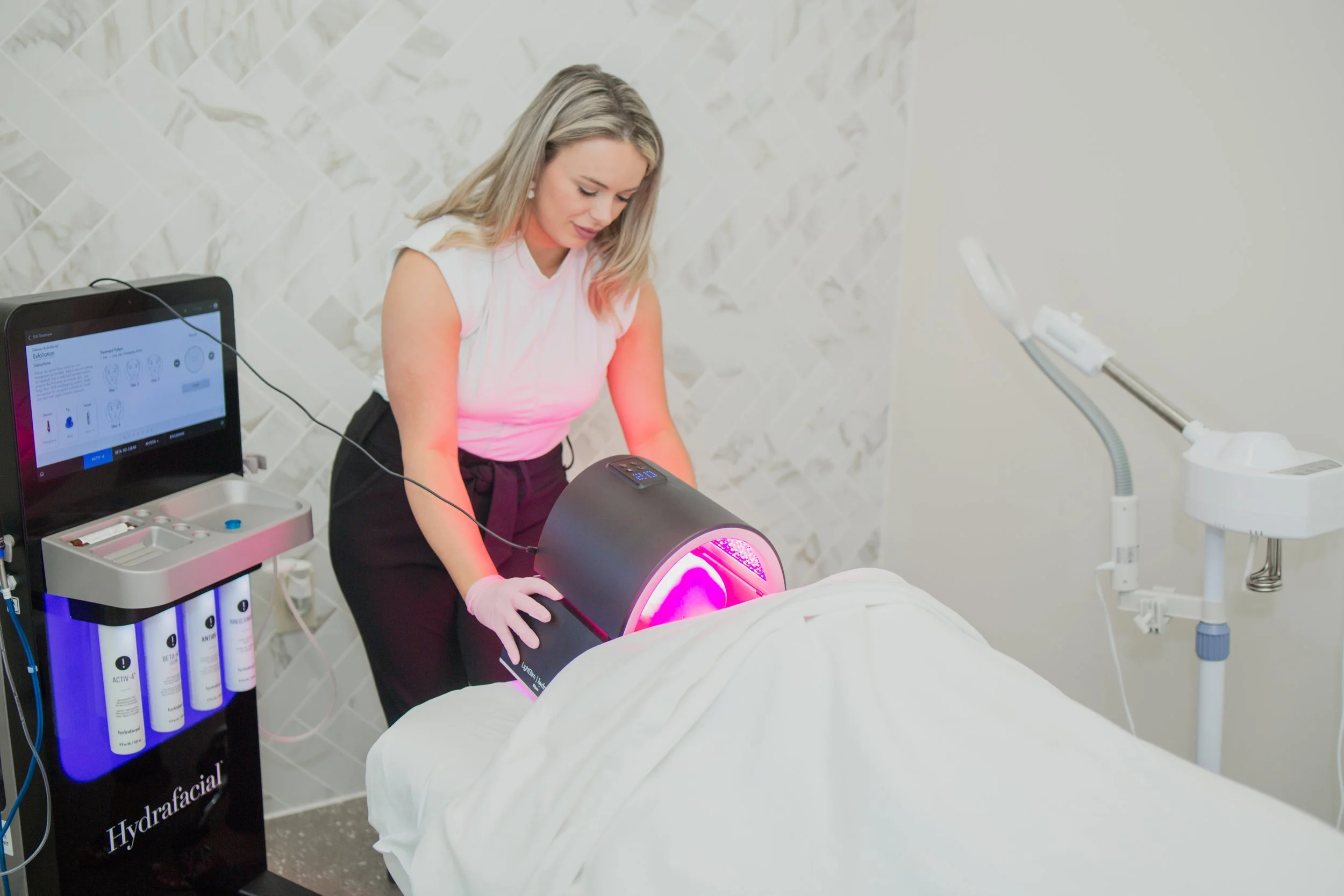 Aesthetician performing LED light therapy on a client using a HydraFacial machine in a spa setting.