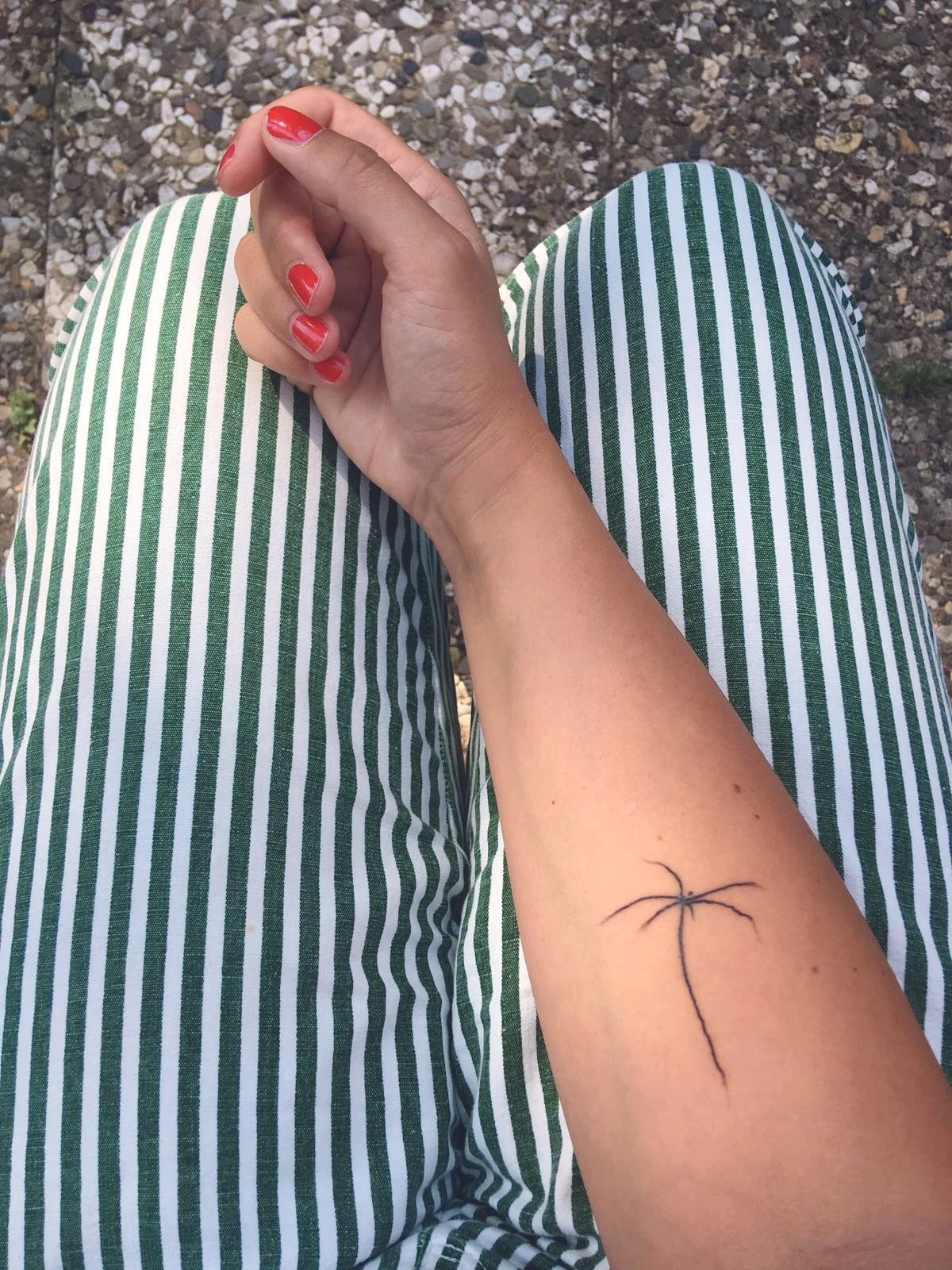 Der Blick von oben auf die Knie einer Person, die grün-weiß gestreifte Hosen trägt, eine tätowierte Unterarmin und rote Nagellack hat. Das Bild ist im Freien auf kiesigem Boden aufgenommen.