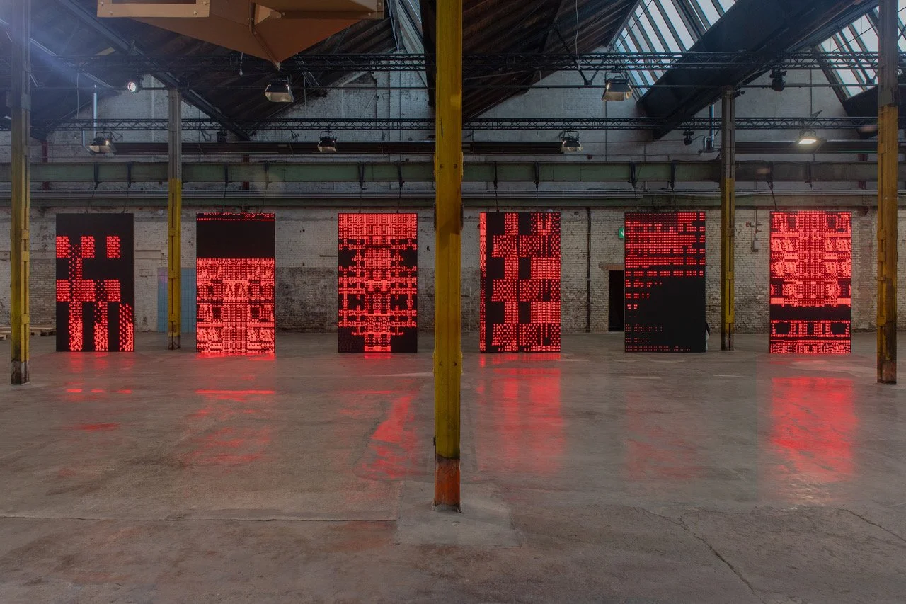 Eine Person geht durch ein Einkaufszentrum oder eine große Halle mit roten LED-Bildschirmen im Hintergrund, die komplexe geometrische Muster zeigen.