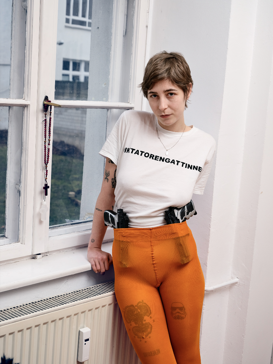 Junge Frau mit kurzer brauner Irofrisur, weißen T-Shirt mit schwarzem Text, Orange Leggings, zwei Pistolen im Hosenbund, steht an einem Fenster und blickt ernst.