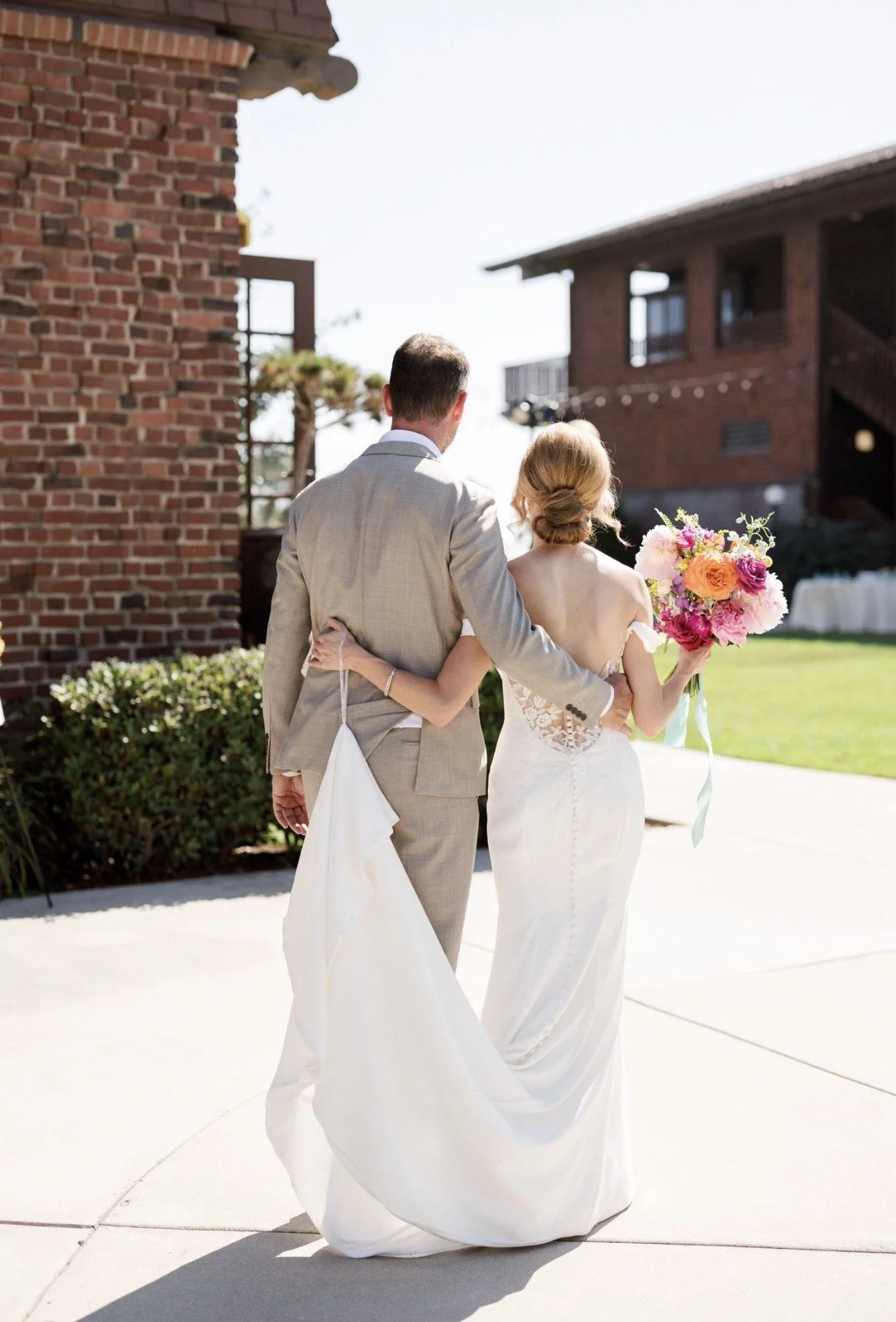 Bride and groom are side by side with their backs facing the camera and their arms wrapped around each other. Bride carries large bright and colorful wedding bouquet.
