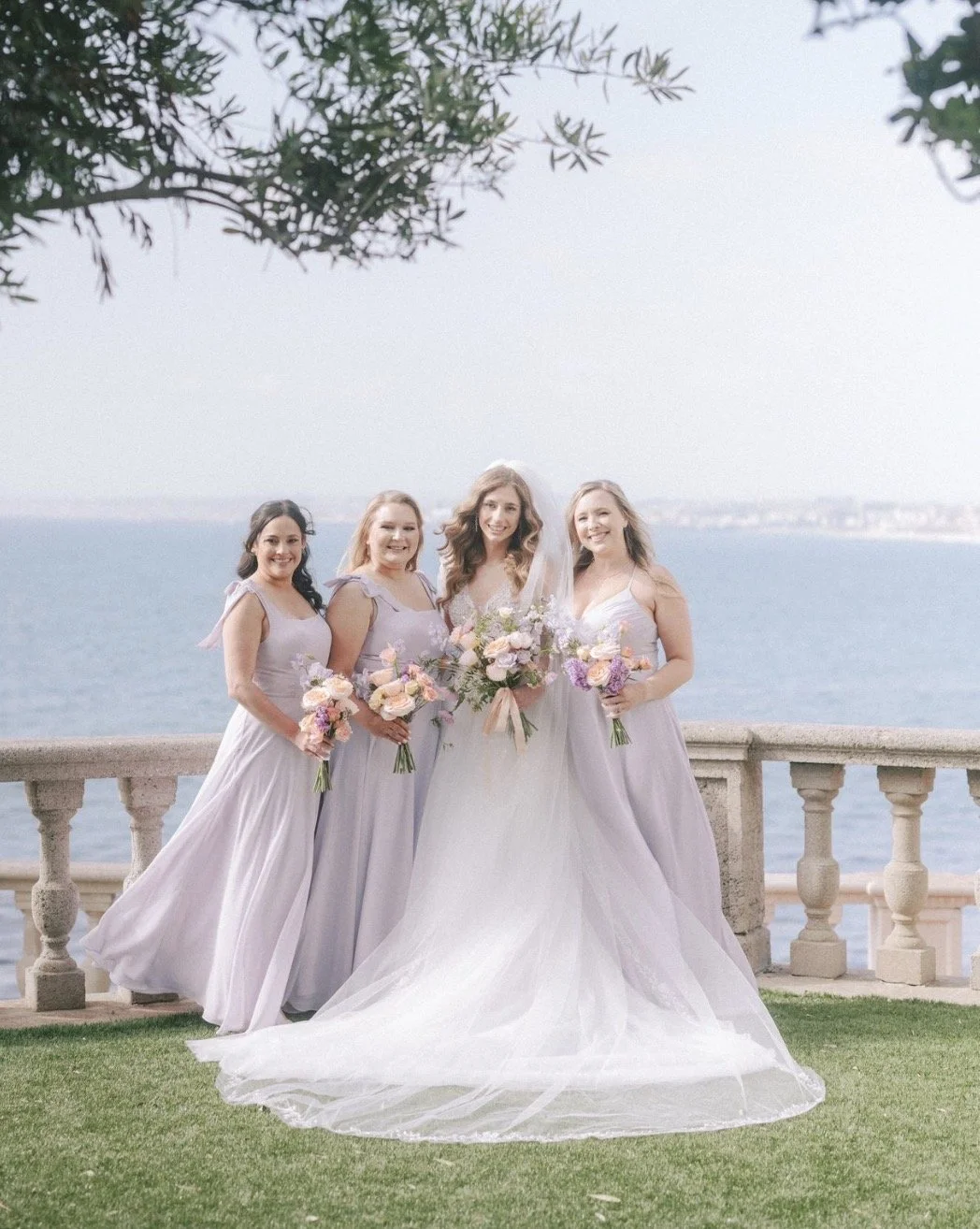 bride and her bridesmaids smiling and standing looking at the camera with the ocean behind them. bride is in white lace and bridesmaids are in light lavender dresses. All carrying floral bouquets of peach and lavender florals