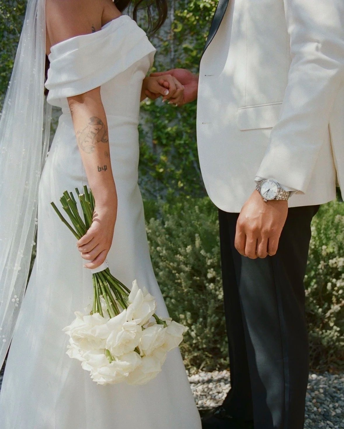 A bride holding a bouquet of white flowers, with an officiant or groom holding her hand, outdoors with greenery in the background.