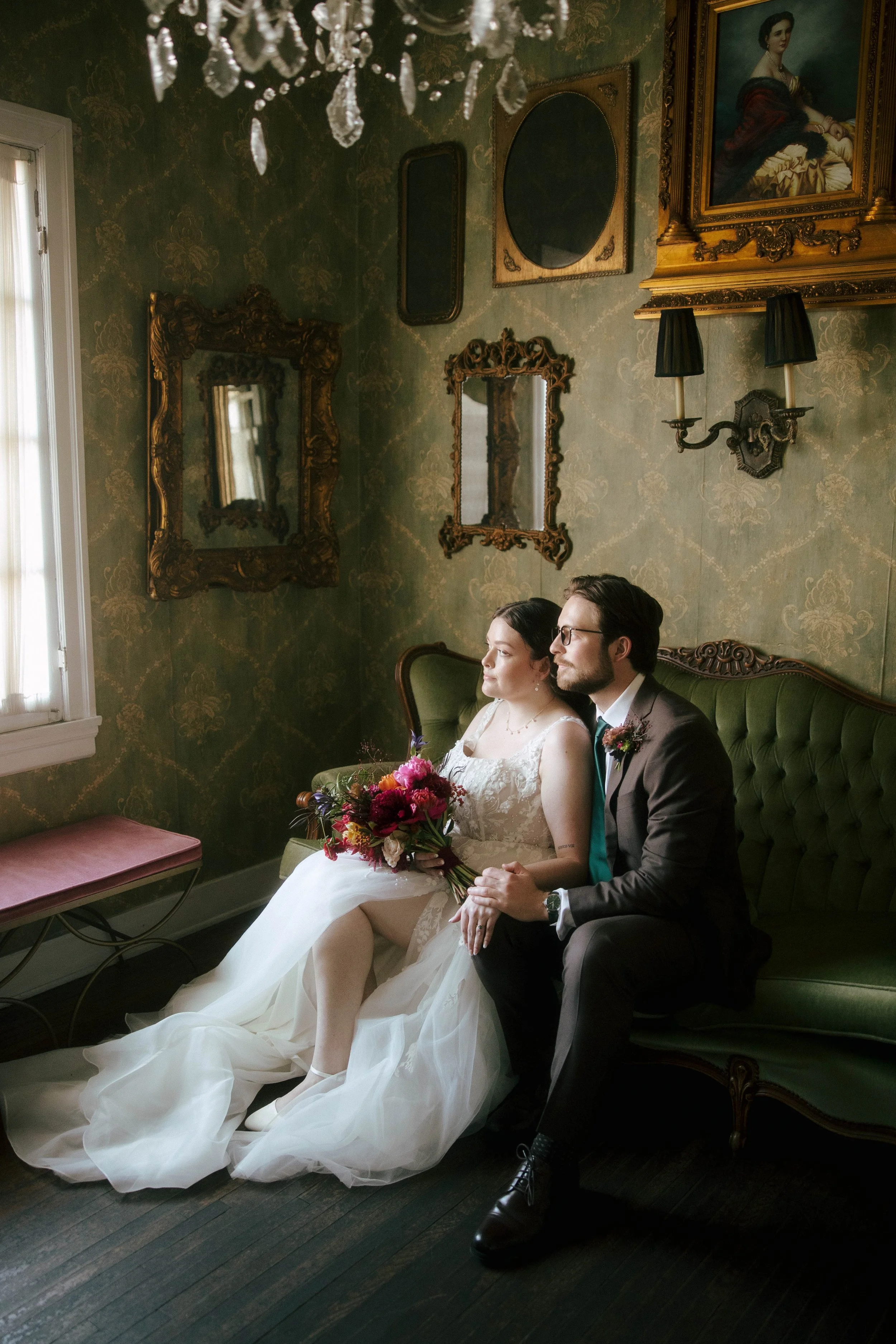 Couple looking out the window in a room with paintings and brass mirrors on the wall. bride in white dress holding jewel colored wedding bouquet. Groom in brown suit with green tie.
