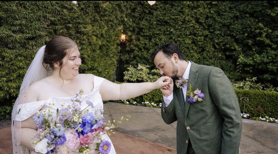 Groom with grey suit and colorful floral pocket square kisses brides outstretched hand. Bride carries a pastel multicolored bridal bouquet and smiles at the groom lovingly.