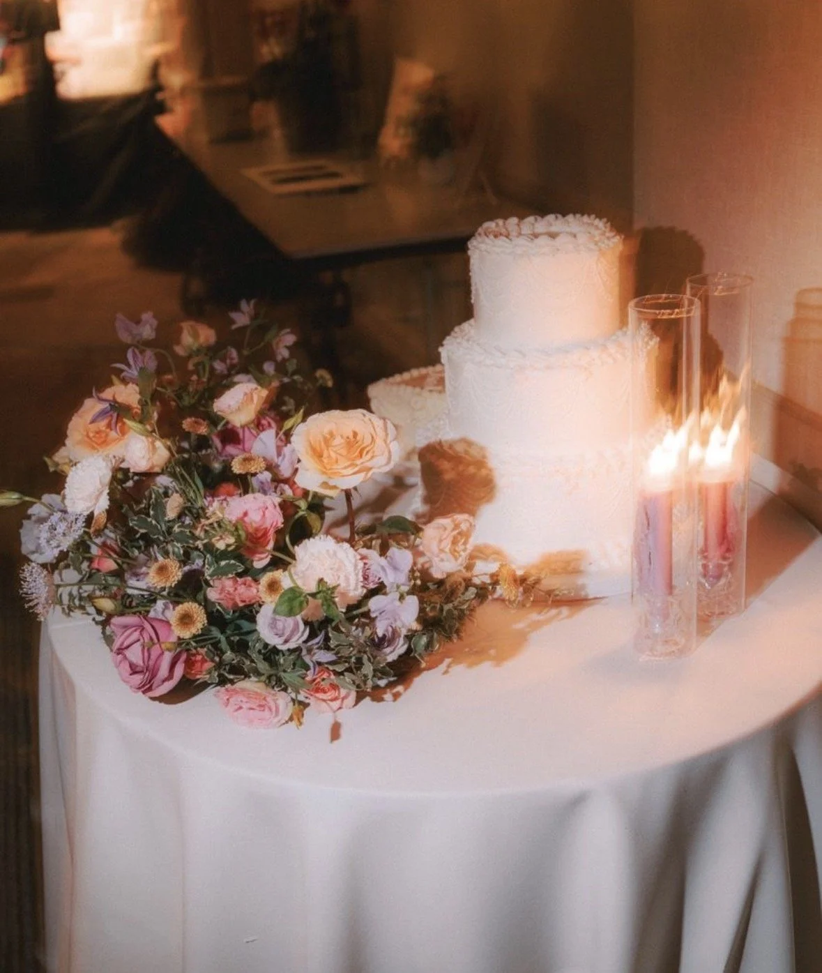 Small circular table with white wedding cake, two candles, and a floral arrangement in peach, white, and lavender florals