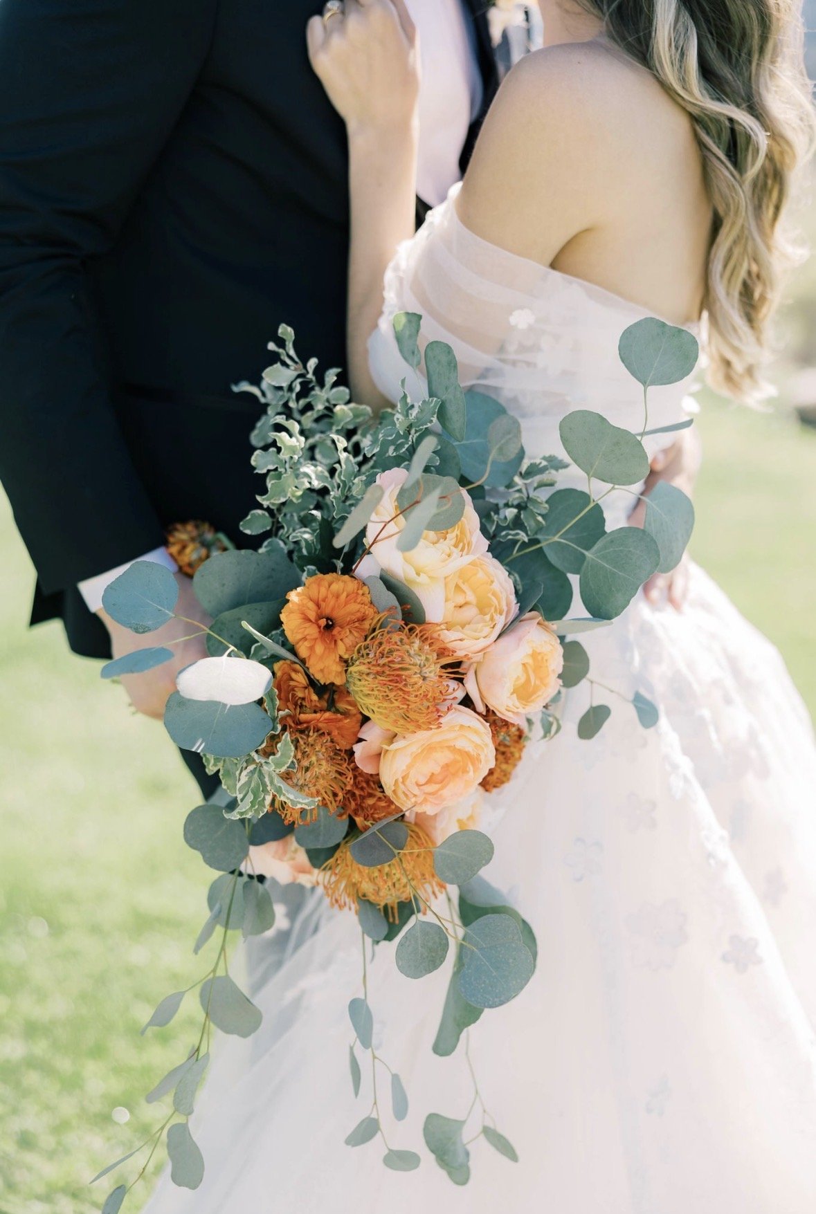 Side view of bride and groom from shoulders to knees. Bride is pressed again groom as groom holds her bouquet facing the camera. Bouquet is made of orange and peach florals and green eucalyptus.