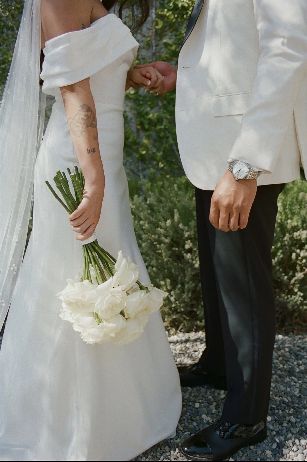 side view of bride and groom facing each other. Next to knee visual only. Grooms watch is visible as well as bride's long stemmed white rose bouquet