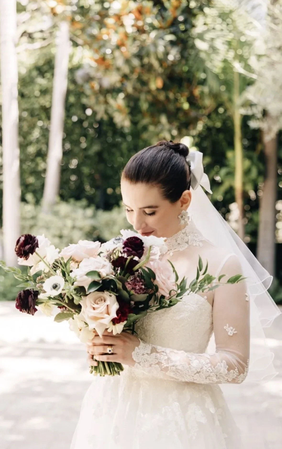 Bride in lace dress smiling down at her large floral bouquet of white, blush, and burgundy florals.