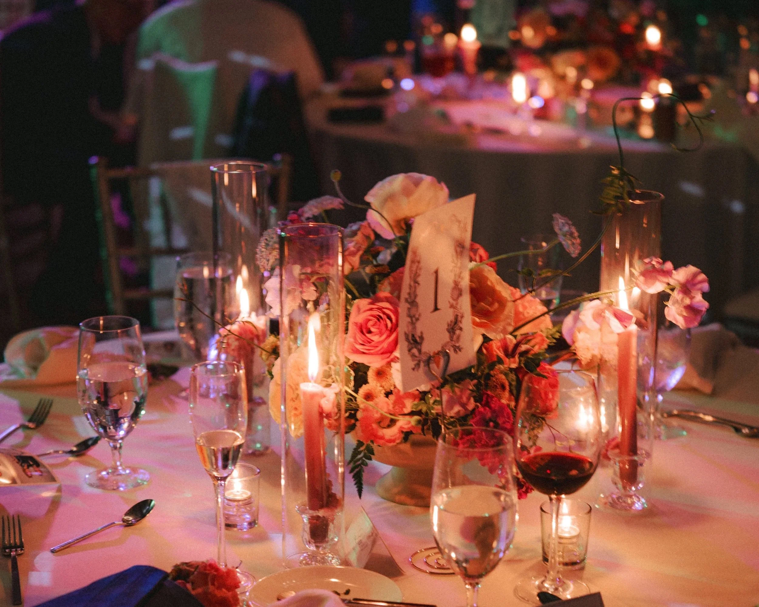 A beautifully decorated dinner table with a floral centerpiece, candles, a wine glass with red wine, glasses of water, and table number 1 in an elegant setting, likely at a wedding or special event.