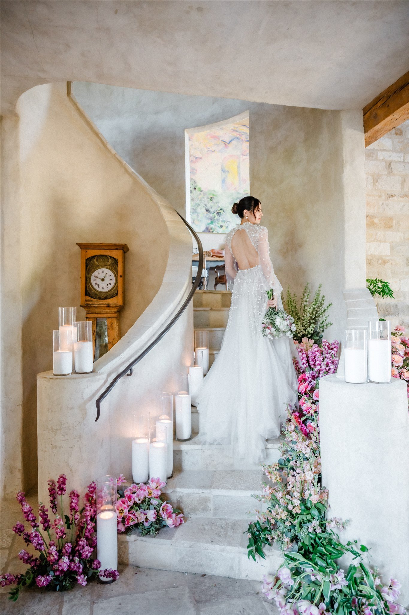 Bride in a wedding gown holding a bouquet, standing on a staircase with pink and white flowers, candles, vintage clock, and artwork in the background.