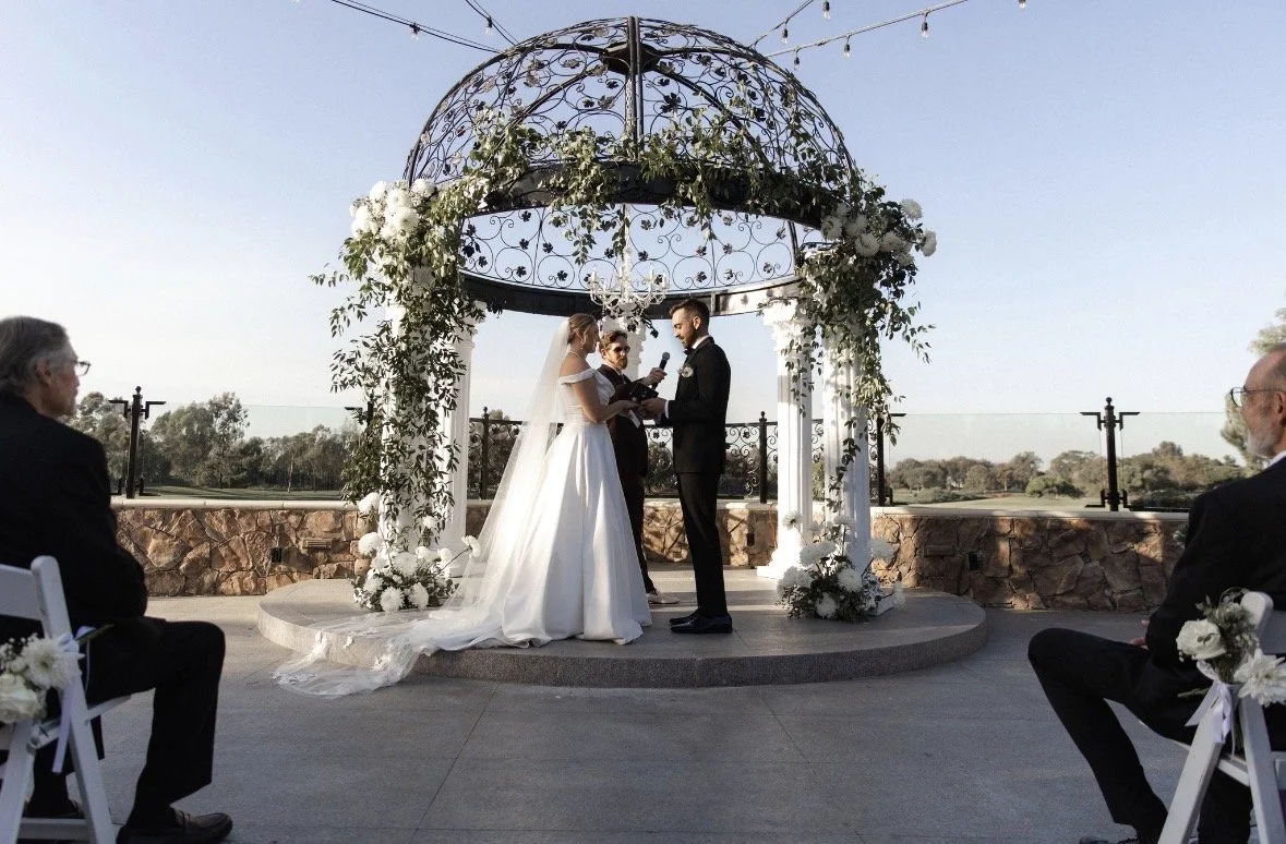 Bride and groom under a gazebo saying their vows. Gazebo is white and iron with white florals and draping greenery.