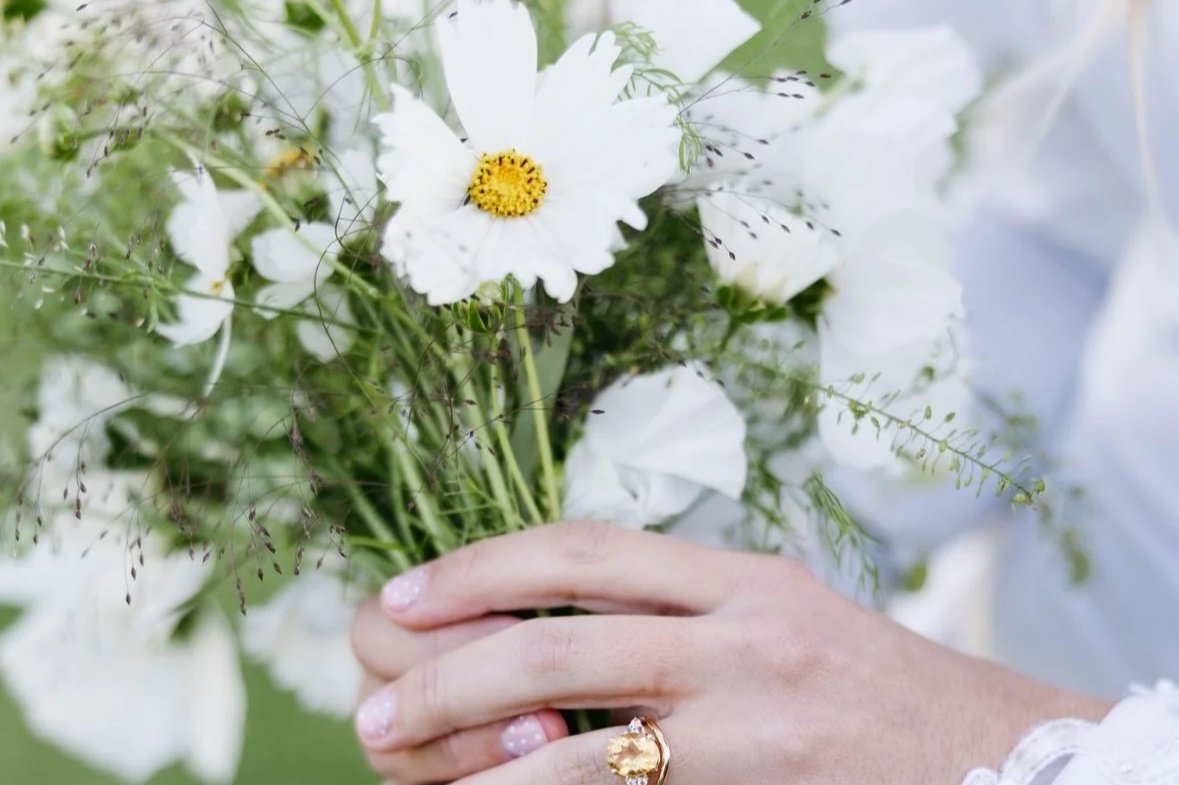 A person holding a bouquet of white flowers with green foliage and a gold ring with a large gemstone on their finger.