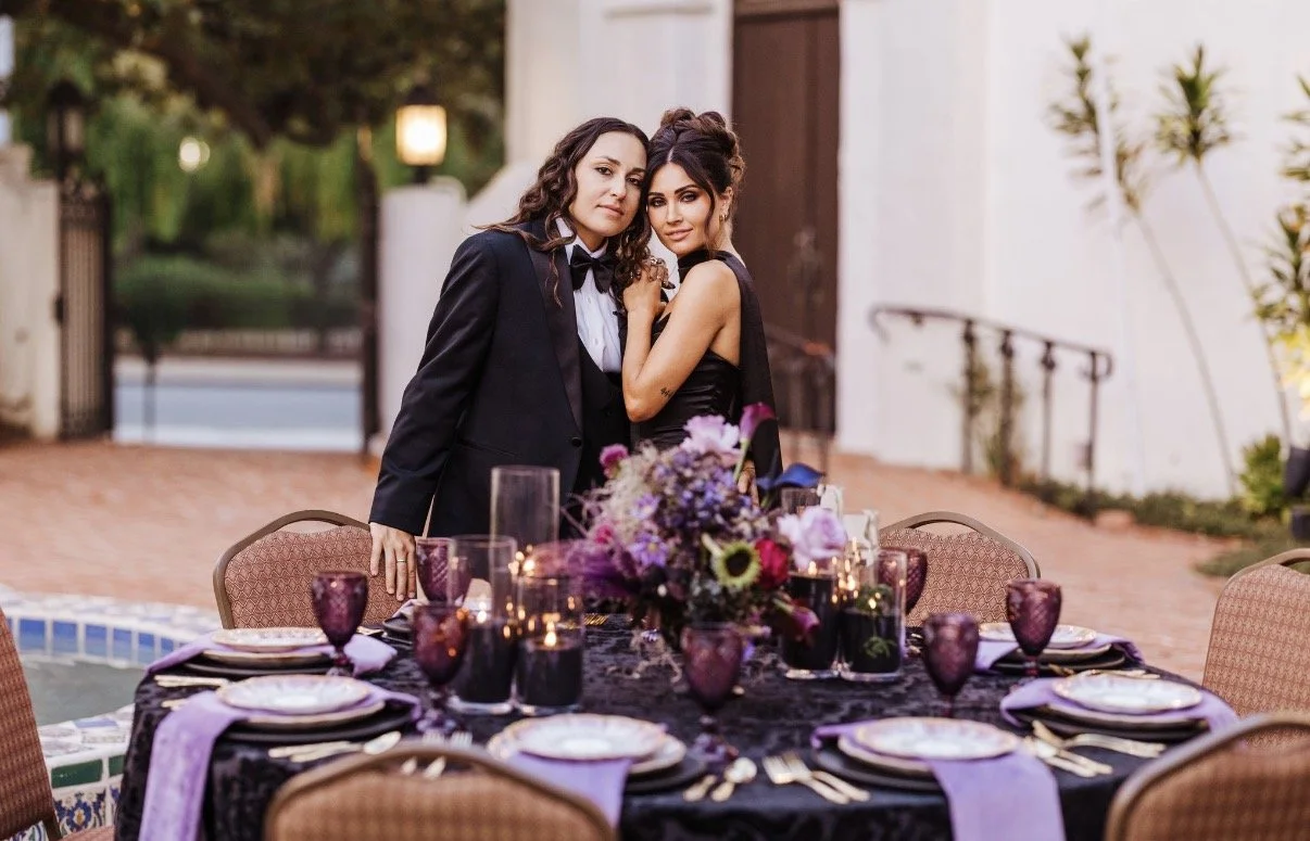 LGBTQ couple both wears black as they stand together facing the camera behind a table at their reception. The table has black and purple linens, black candles, and a purple floral centerpiece.