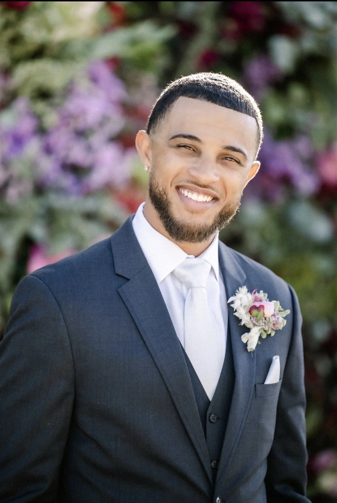 Smiling groom looking at camera. Her has dark skin and a well trimmed short beard, and a diamond earring. he is wearing a dark greyish blue suit and a fluffy pink and white boutonnière.