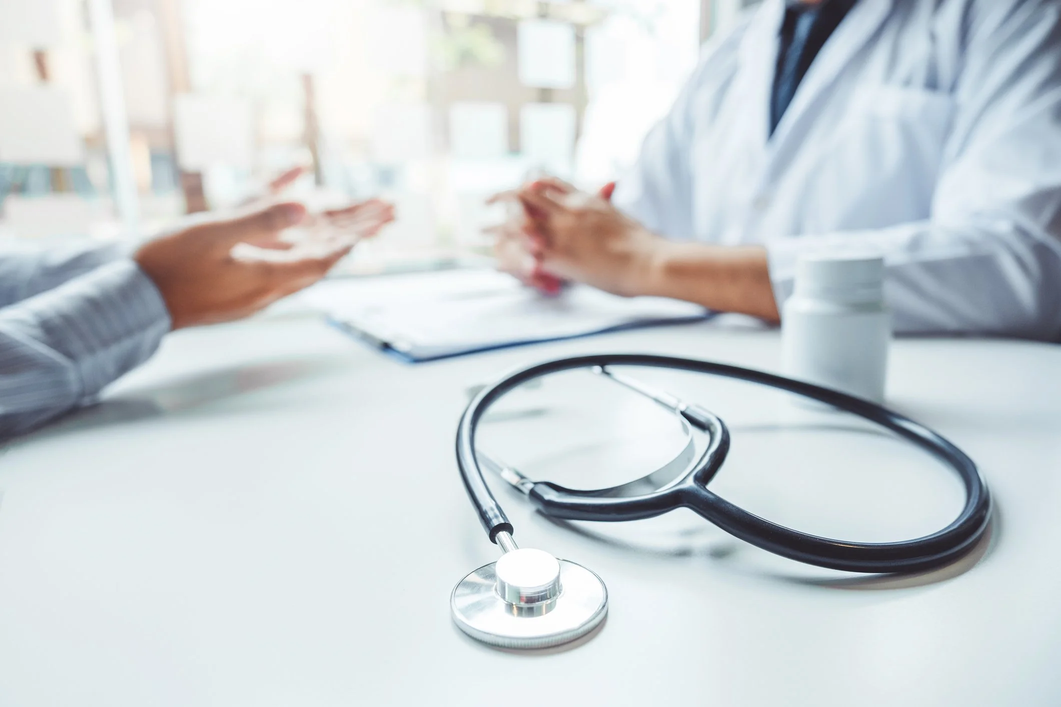 A stethoscope on a white desk with healthcare professionals in the background during a consultation.